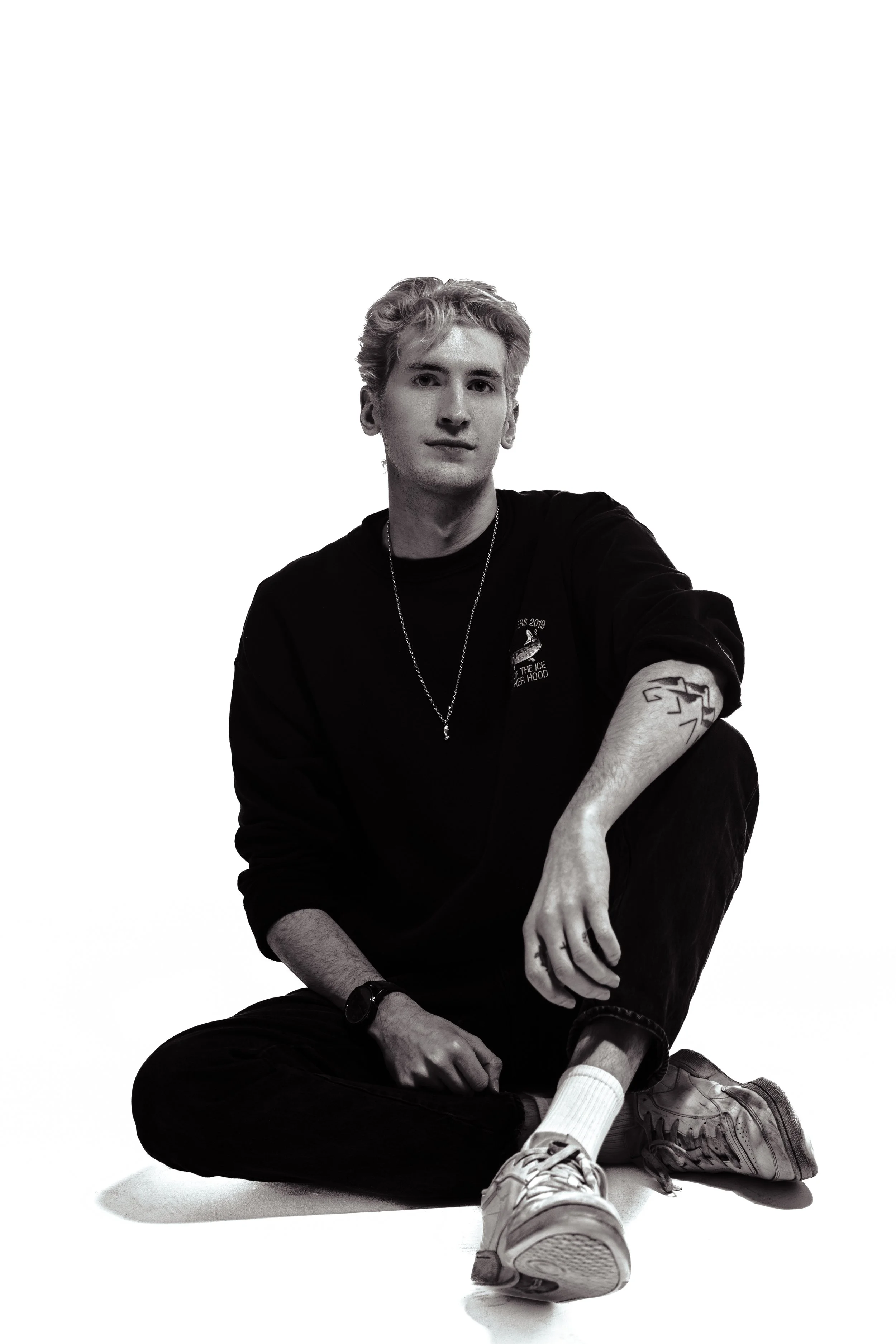 Black and white photo of Hunter Cairns with light hair, sitting on the floor with one knee up. He's wearing a black shirt, black pants, sneakers, and a watch, with a chain necklace. His hair is slightly tousled, and he has a tattoo on his arm.