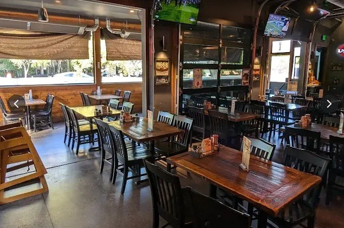 Interior of a casual restaurant with wooden tables and black chairs, TV screens on the walls, and large windows letting in natural light.