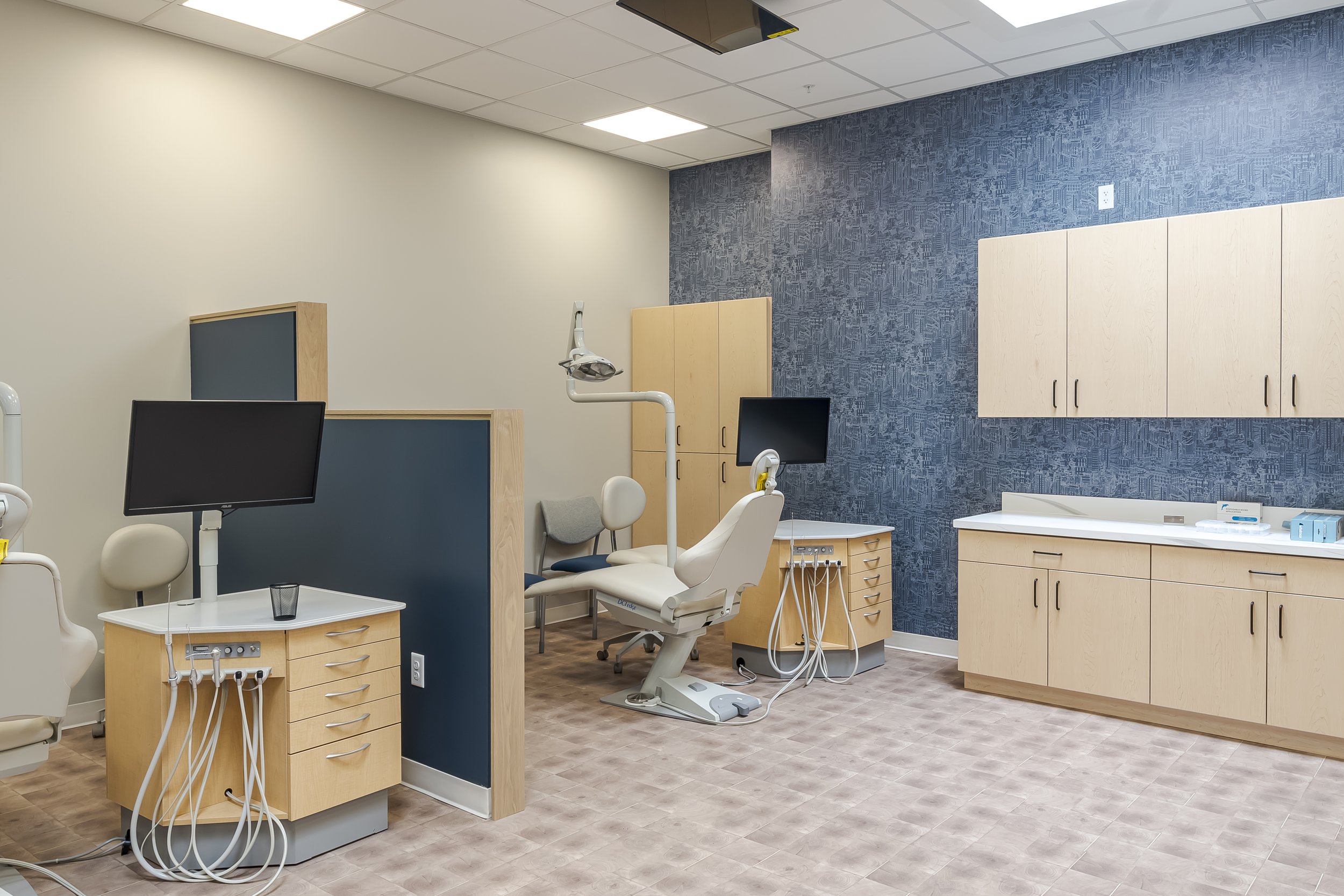 A dental clinic examination room with dental chairs, monitors, and storage cabinets, featuring a blue accent wall and ceiling lighting.