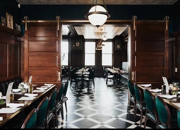 Elegant restaurant interior with dark wood paneling, green upholstered chairs, set tables with white napkins and flowers, and large windows letting in natural light.