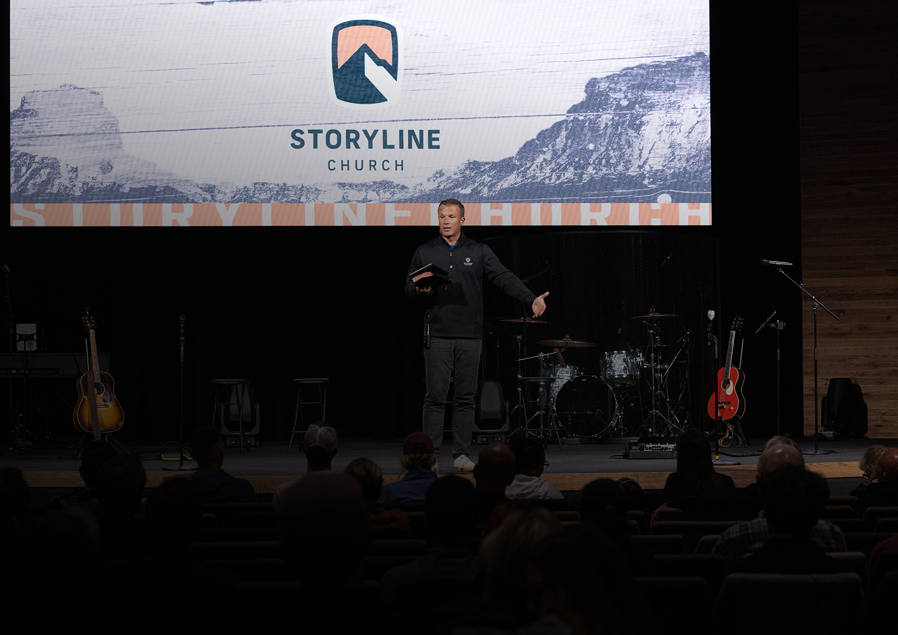 A man stands on a stage speaking to an audience in a church, with a large screen behind him displaying the logo and name of Storyline Church. The stage has musical instruments including guitars and drums, and the audience is seated in front of him.