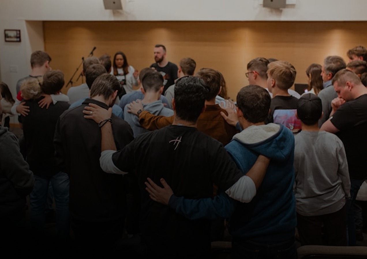 A group of people, mostly young adults, standing closely together with their arms around each other's shoulders in prayer or reflection, facing towards the front of the room where two individuals are standing at a distance.