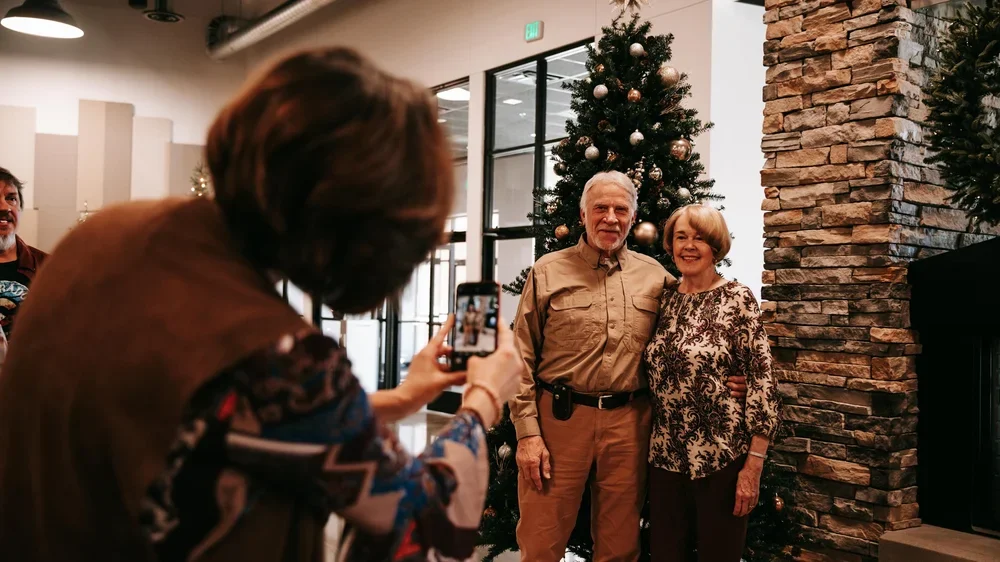 A woman taking a photo of an elderly man and woman posing in front of decorated Christmas trees inside a modern indoor space.