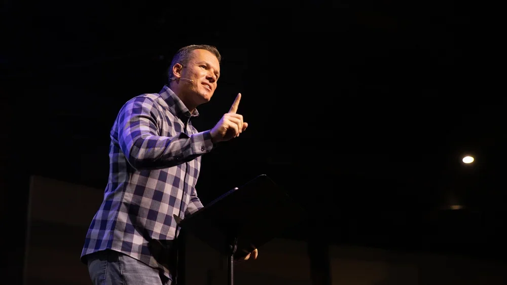 A man standing at a podium on a stage, speaking and gesturing with his right hand, in a dimly lit auditorium or conference room.