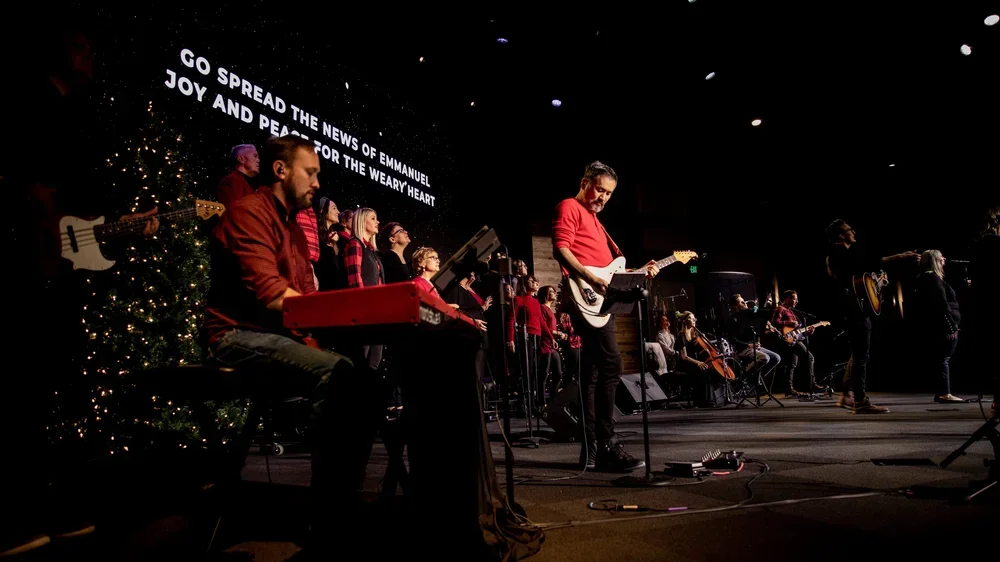 A group of musicians and singers performing on stage with Christmas decorations in the background. A large screen displays a message about spreading the news of Emmanuel from the Bible.