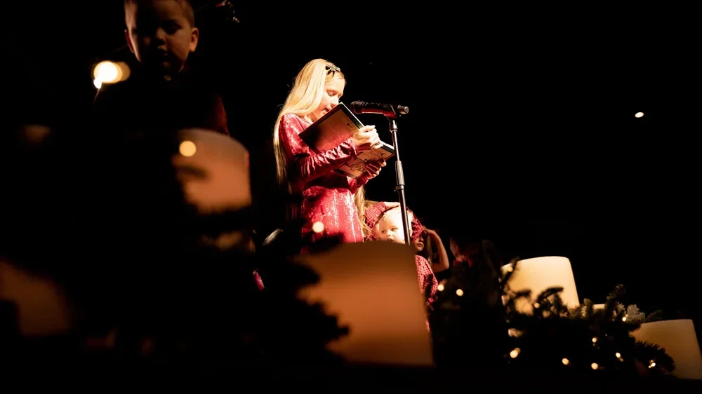 A woman in a red dress reading from a book and speaking into a microphone on stage, with children nearby, dim stage lighting, and Christmas decorations in the foreground.