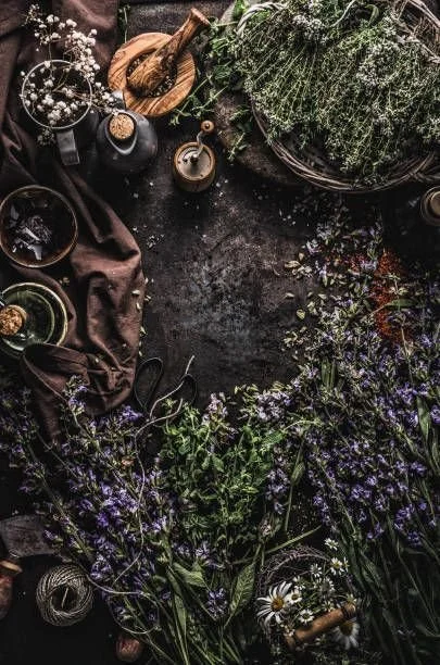 A dark rustic table with various flowers, herbs, and gardening supplies arranged around it, including a basket of herbs, a small wooden cutting board, a brown cloth, and containers with decorative items.