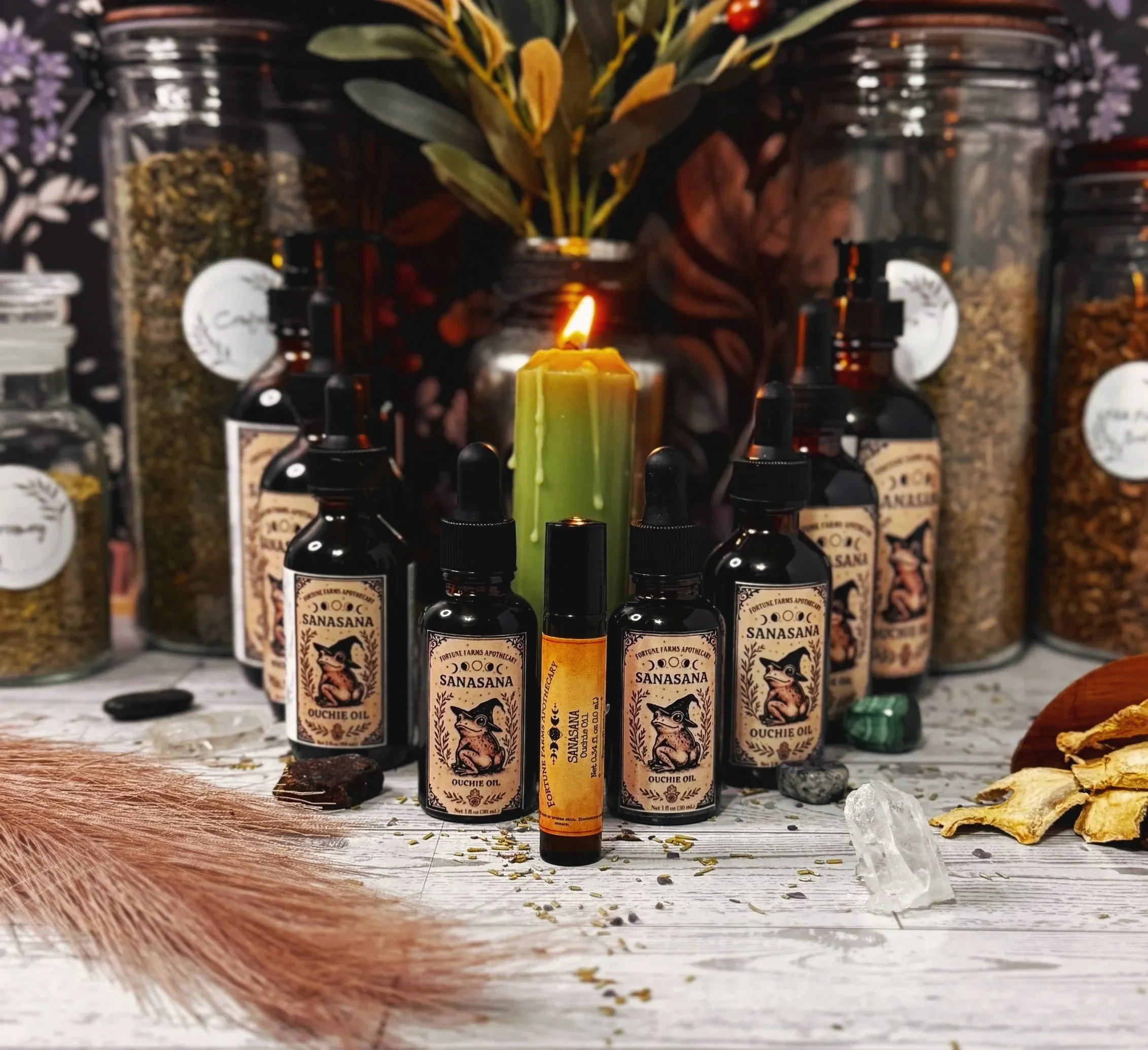 Display of various herbal oils and medicinal items, including a lit green candle, glass jars with dried herbs, and crystals, arranged on a wooden surface with a dark floral background.