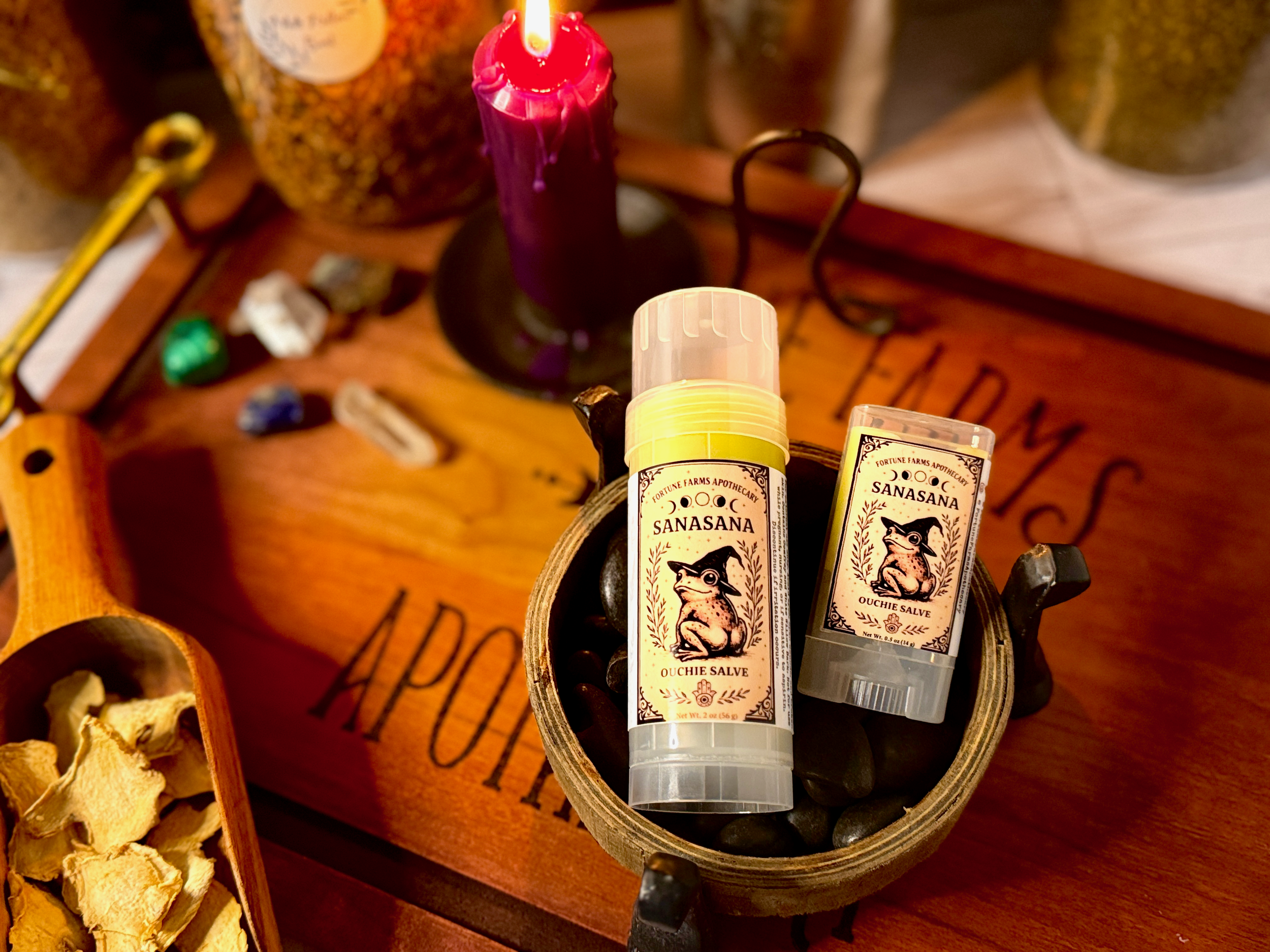 A wooden tray with black stones, a tube and a container of Sanasana Ouchie Salve, a pink candle, and various small decorative items.