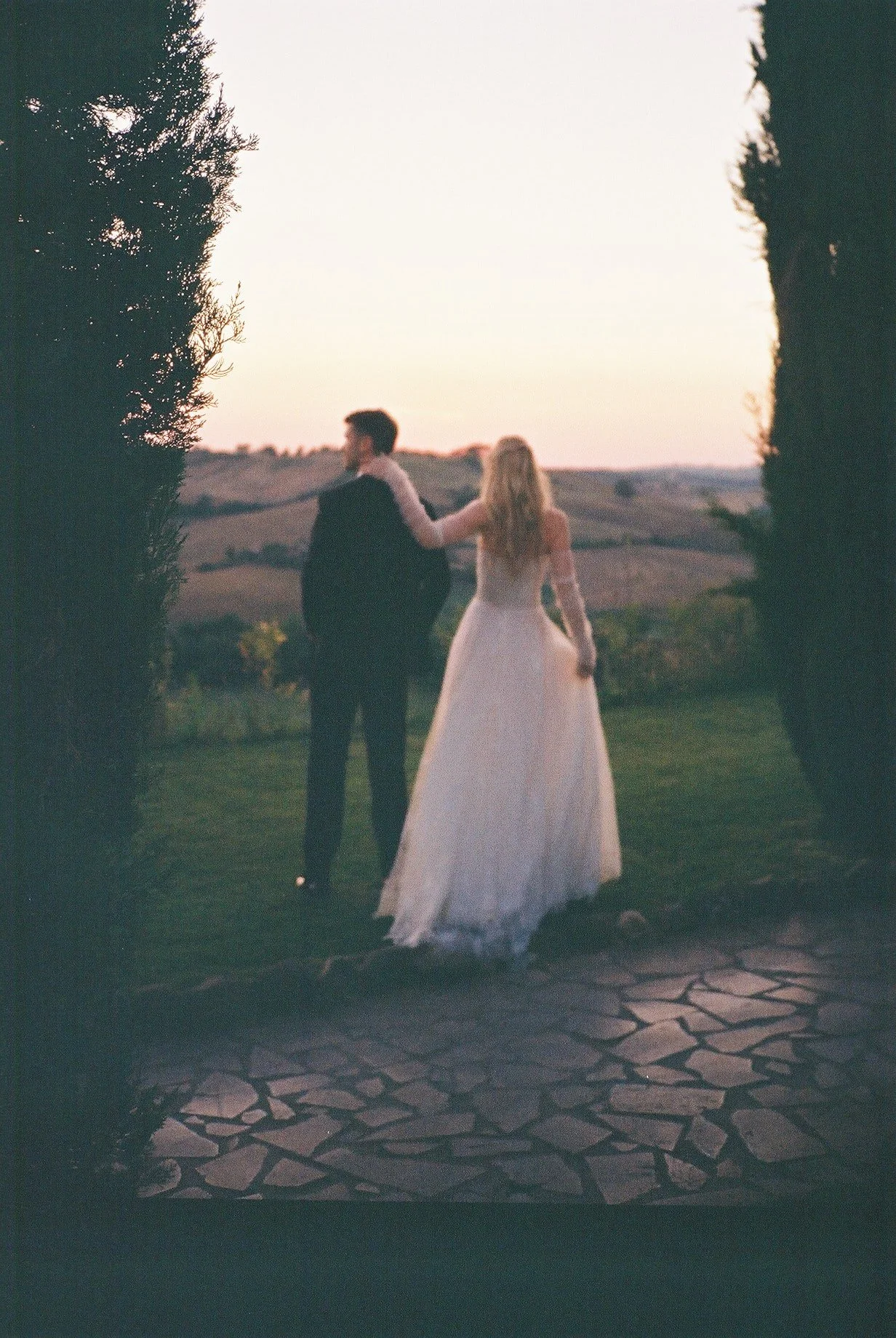 A bride and groom holding hands outdoors at sunset, framed by trees, with hills in the background.