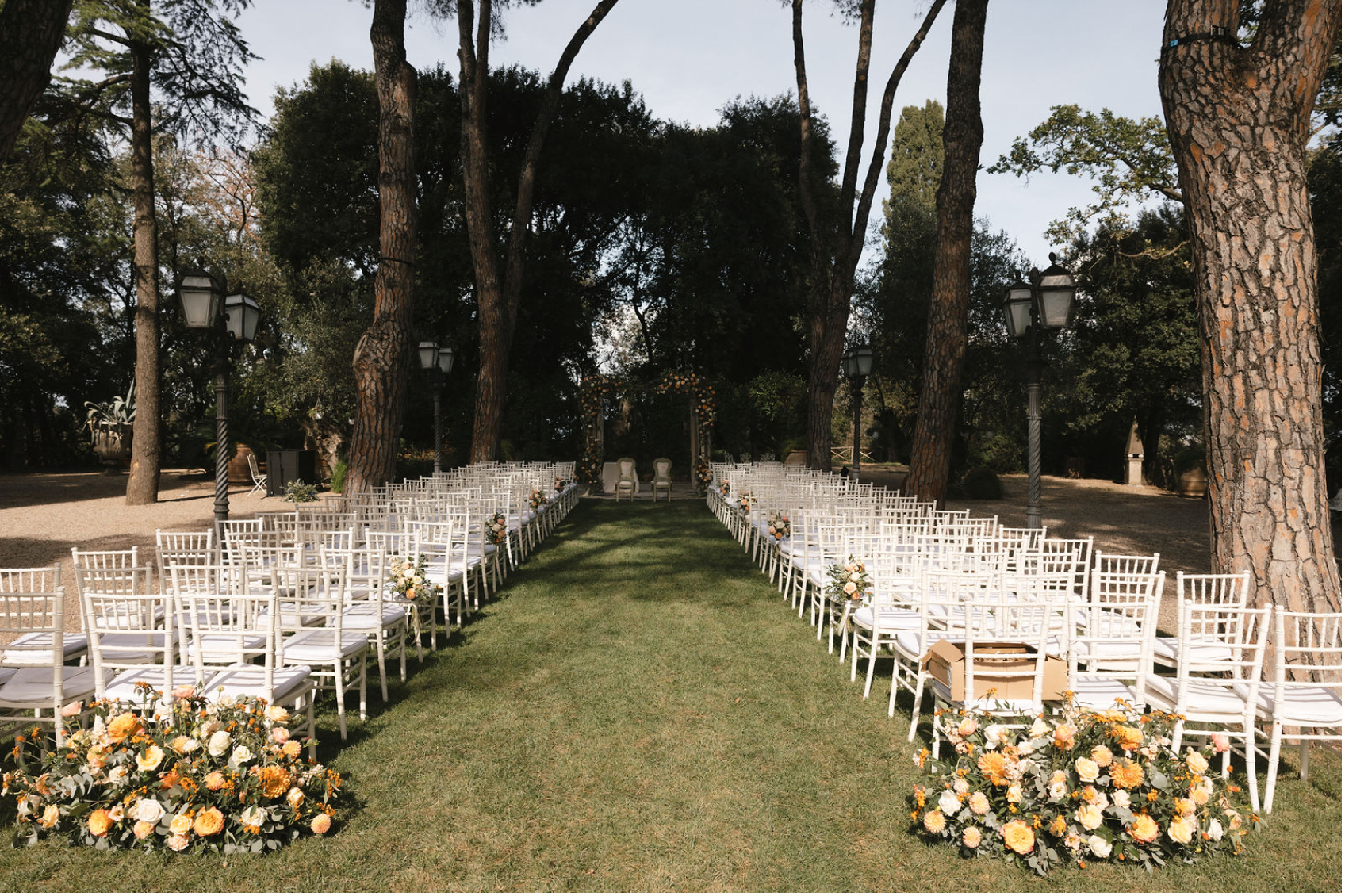 Wedding ceremony setup outdoors with rows of white chairs, floral arrangements, and a decorated arch at the end of the aisle under tall trees.