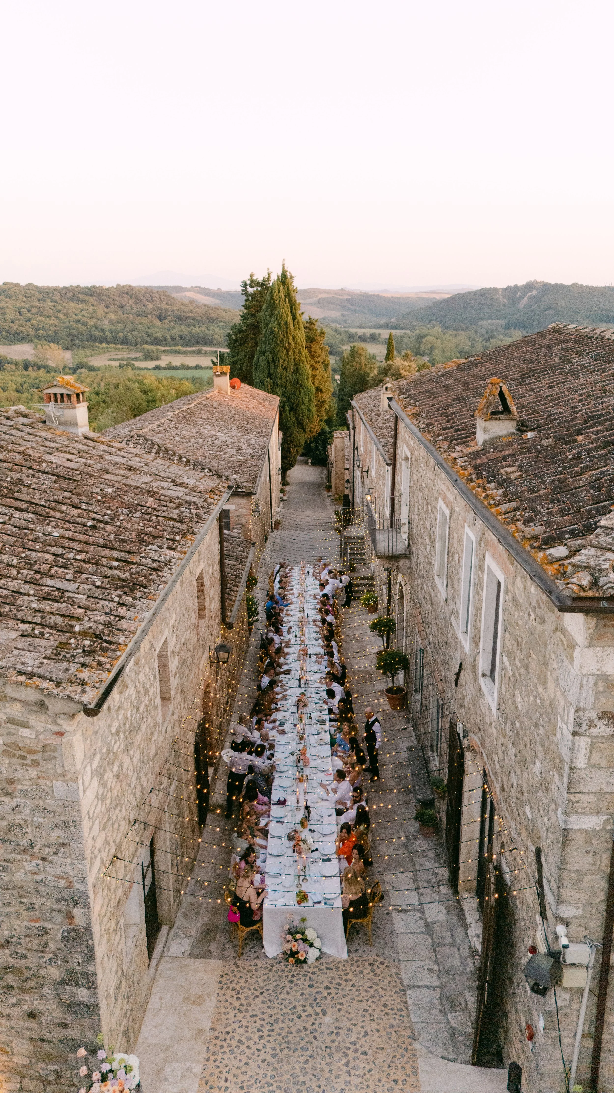 Outdoor wedding reception with a long table set between stone buildings, decorated with string lights and floral arrangements, overlooking a scenic landscape with hills and trees at dusk.