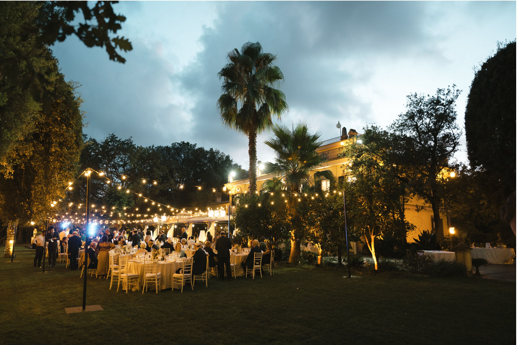 Outdoor evening event with illuminated string lights, round tables with white tablecloths, and guests socializing on a lawn surrounded by trees and a large mansion with warm lighting in the background.