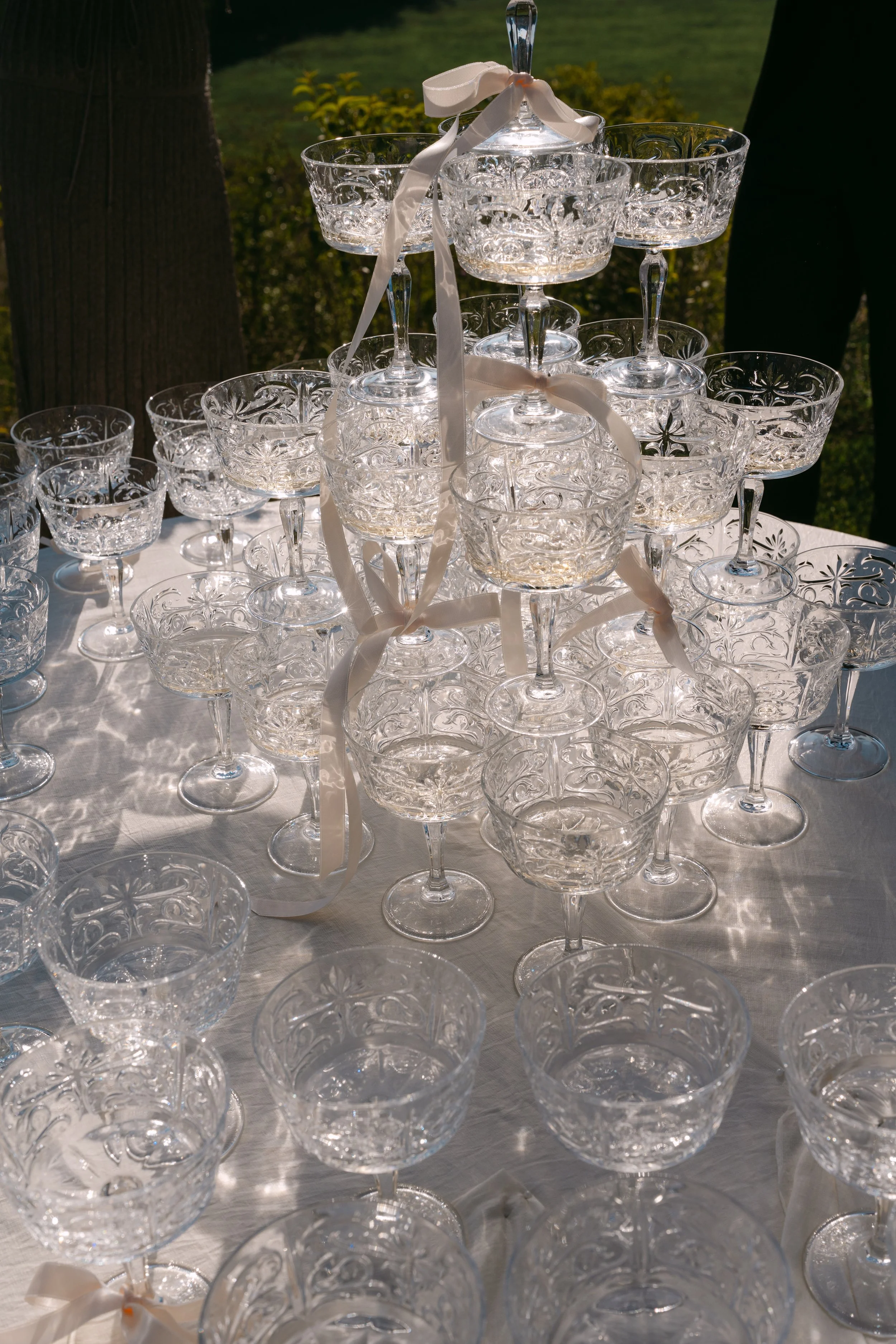 Collection of cut crystal glasses arranged in a pyramid shape on a table at an outdoor event, with sunlight reflecting off them.