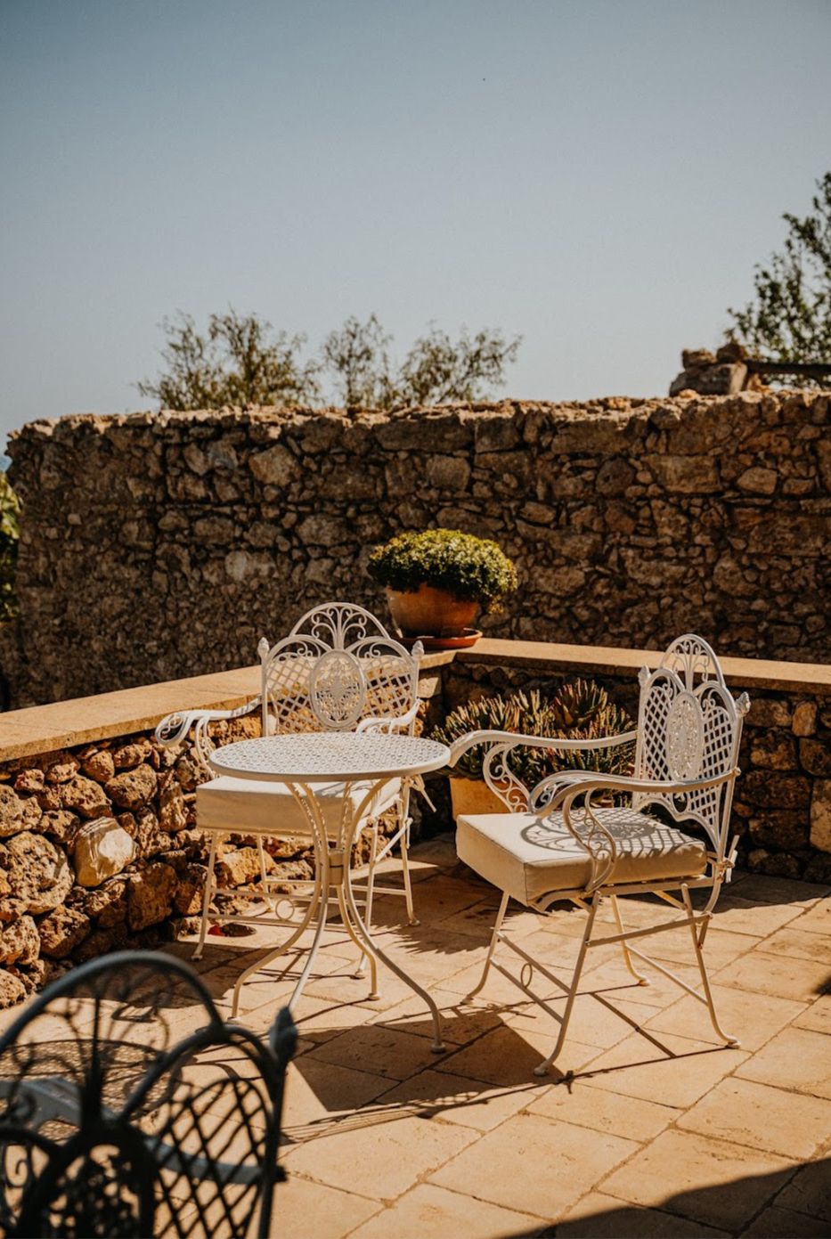 Outdoor patio area with white wrought iron table and chairs, decorative plant pots, a large potted plant, and a stone wall in the background.