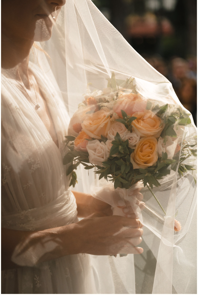 A woman in a wedding dress holding a bouquet of peach, white, and pink roses wrapped in white ribbon, with a delicate veil partially covering her face, on a sunny day.