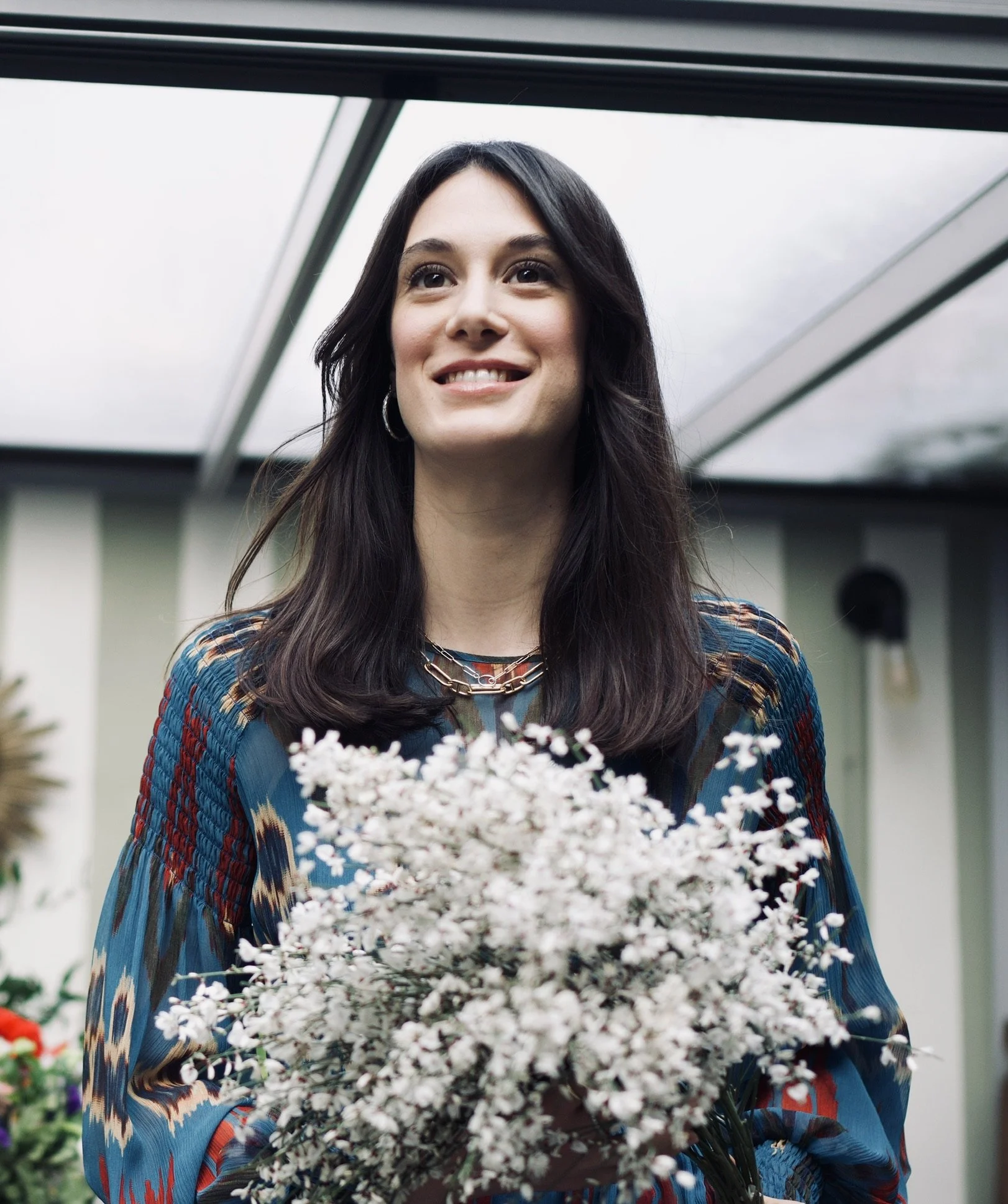 A woman with dark hair holding a bouquet of white flowers, smiling, inside a greenhouse.