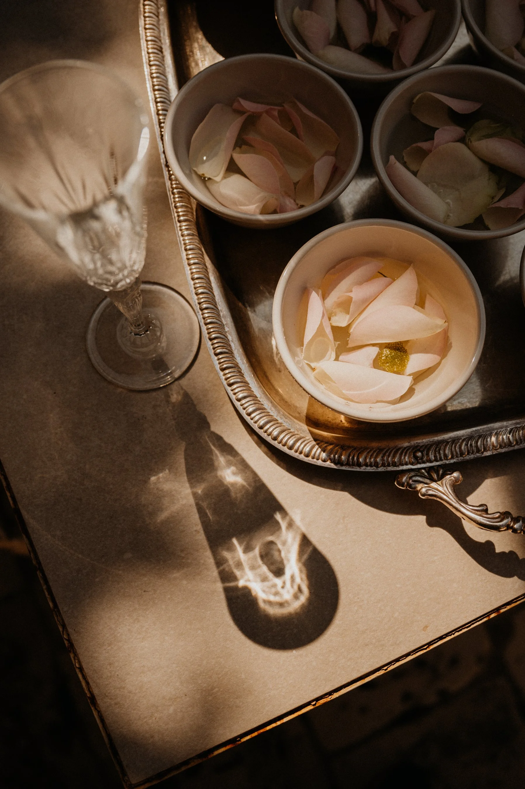 A silver tray with bowls of flower petals and a glass of champagne on a table with sunlight creating shadow patterns.