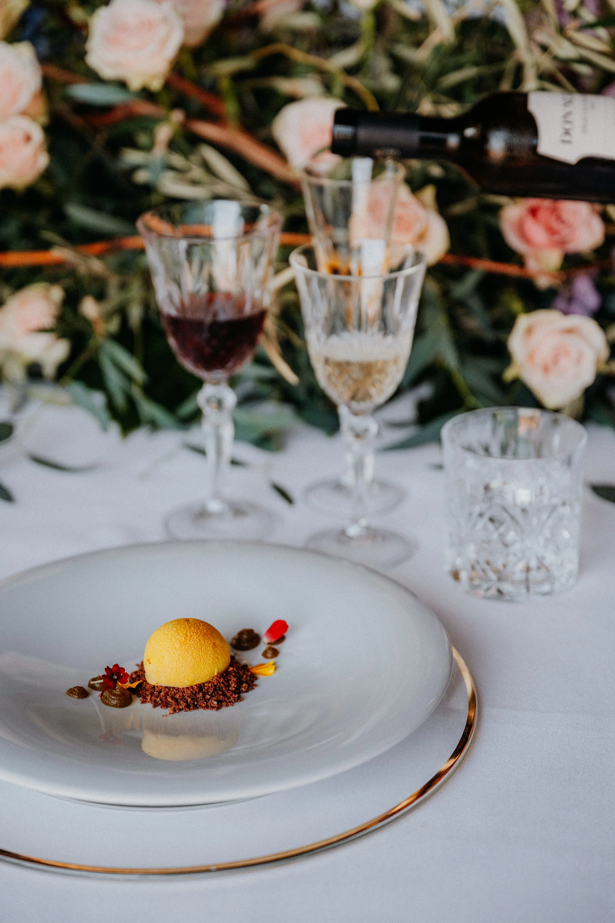 Elegant dessert on a white plate with a yellow scoop, surrounded by small red flower petals and chocolate crumbs, set on a table with glasses of red and white wine and a floral backdrop.