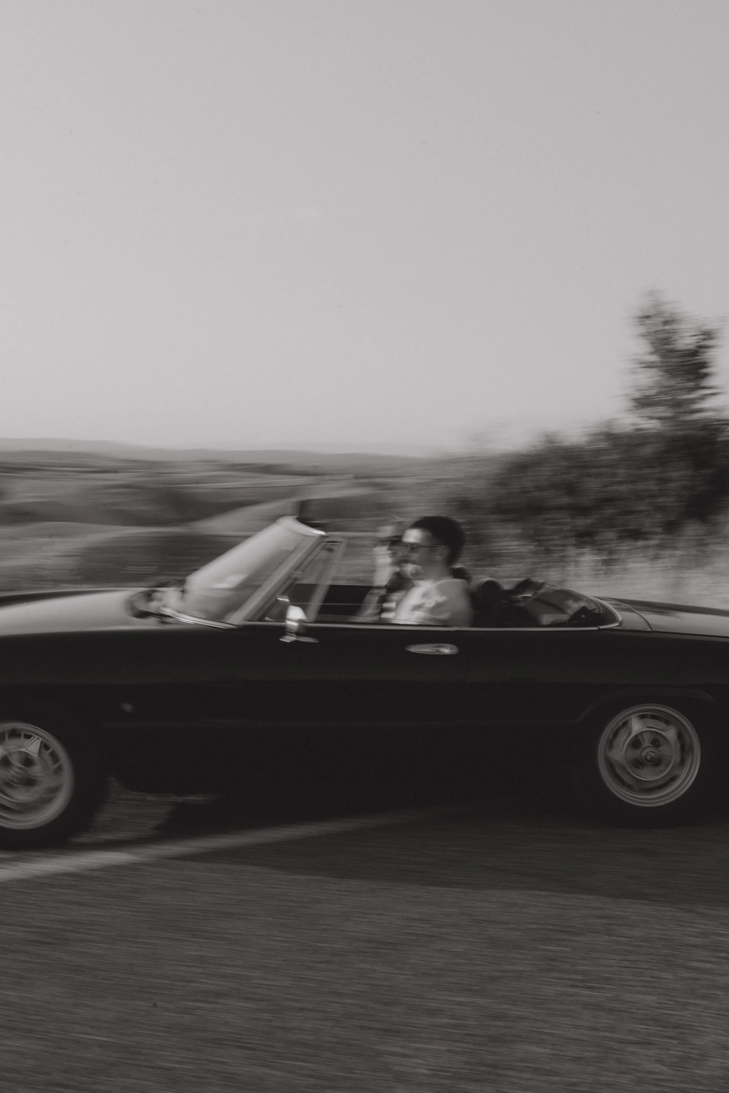 Black vintage convertible car driving on the road with two people inside, one wearing sunglasses, in motion blur, black and white photograph.