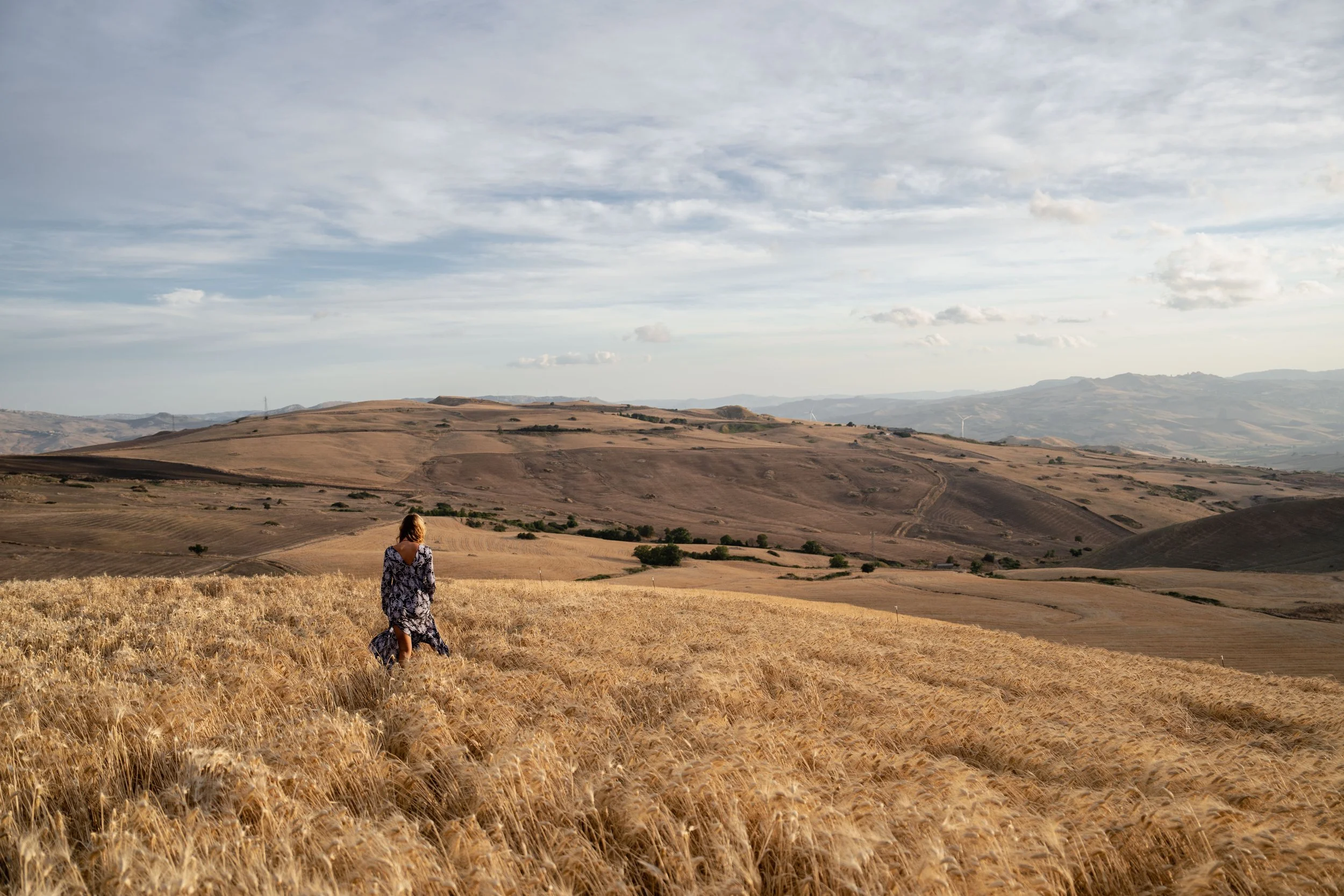 A woman walking through a golden wheat field under a partly cloudy sky, with rolling hills in the background.