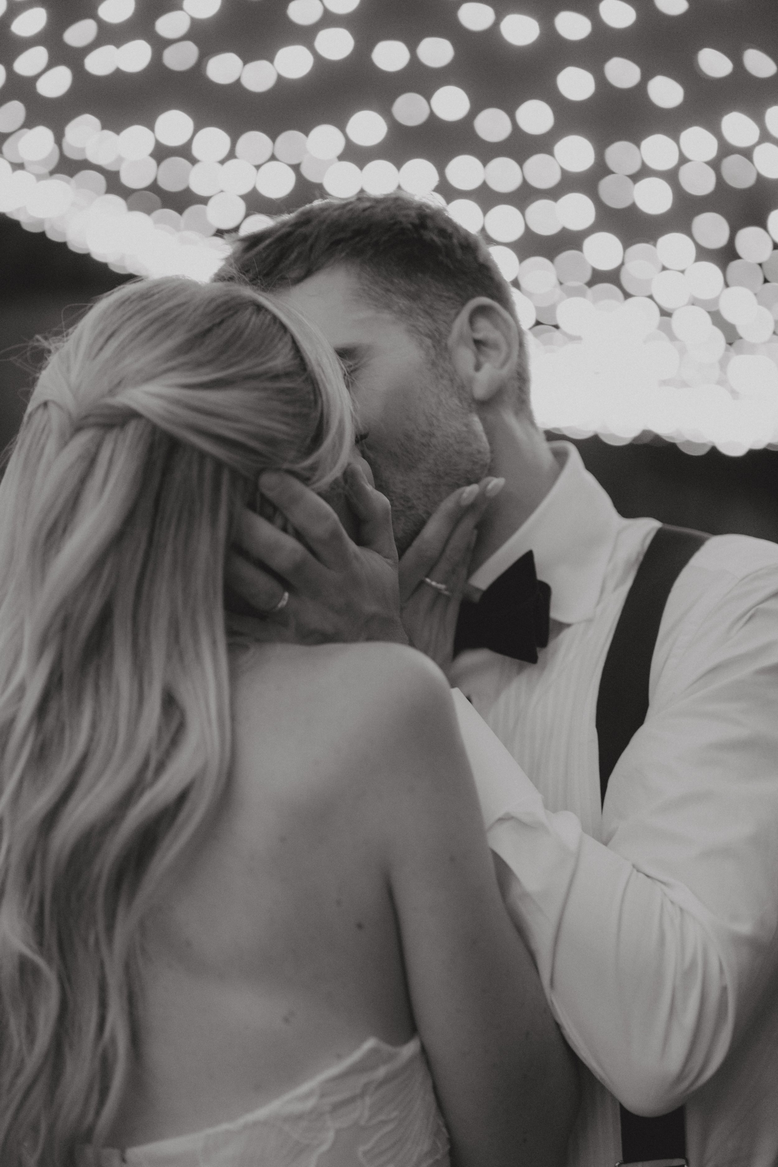 A black and white photo of a couple kissing at a formal event, with string lights in the background.