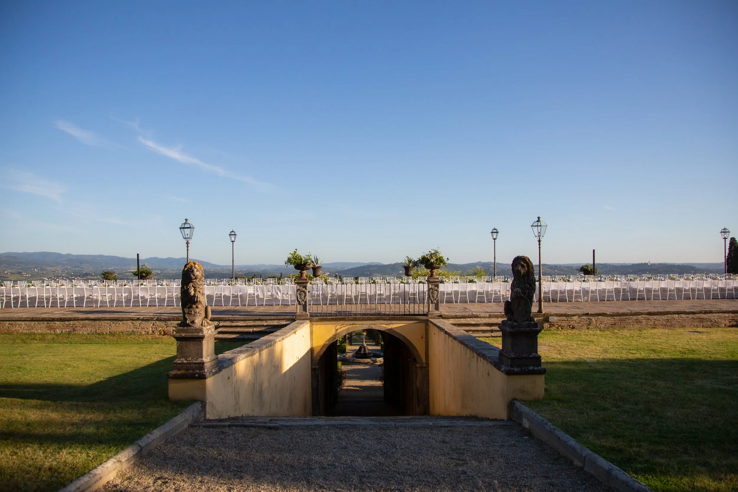 Outdoor banquet setup with tables and chairs on a terrace, decorative statues on pillars, and landscape in the background under a clear blue sky.