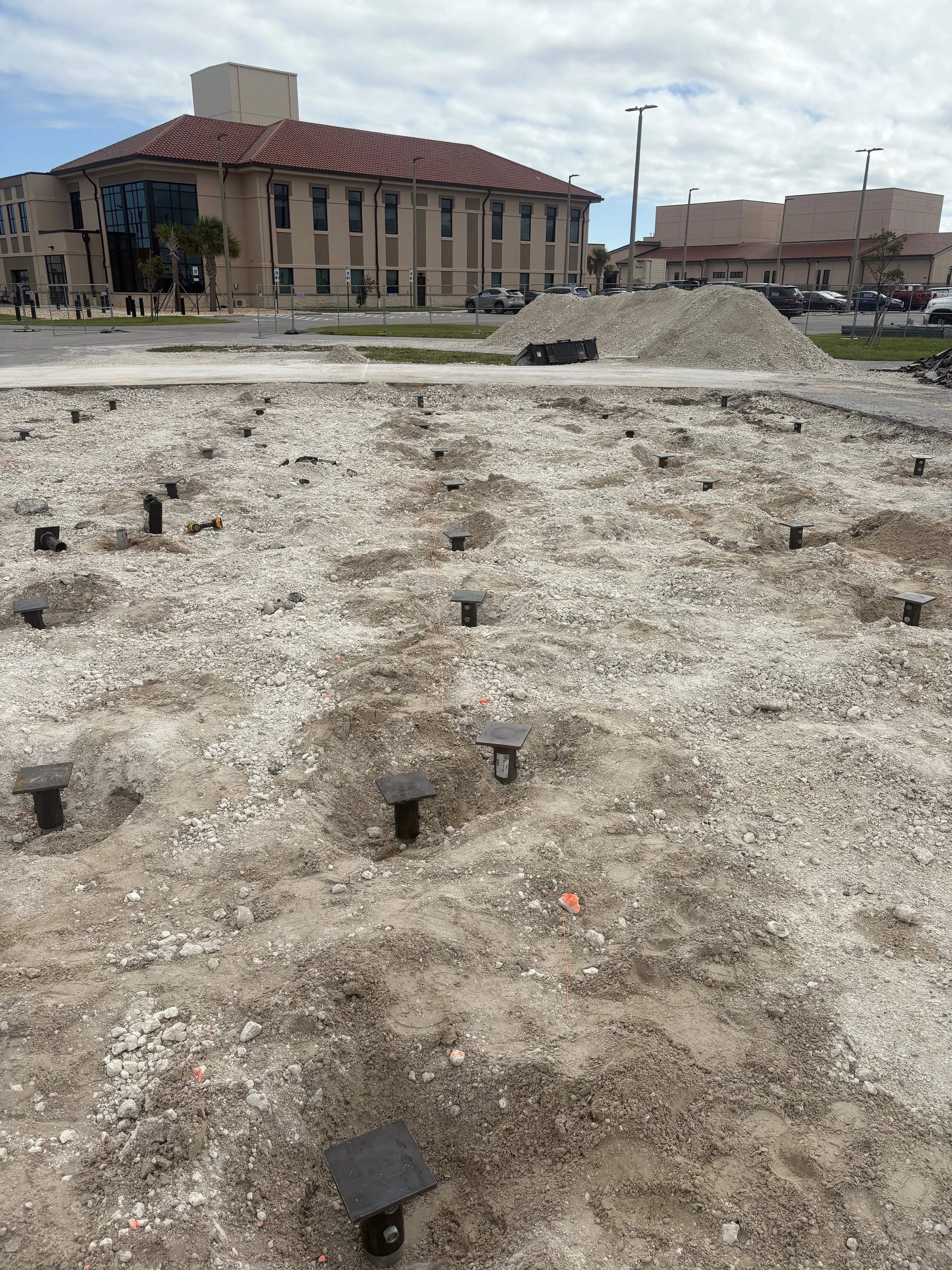 Construction site with exposed ground, small black mounting brackets, a string line, and a pile of gravel, with buildings and parked cars in the background.