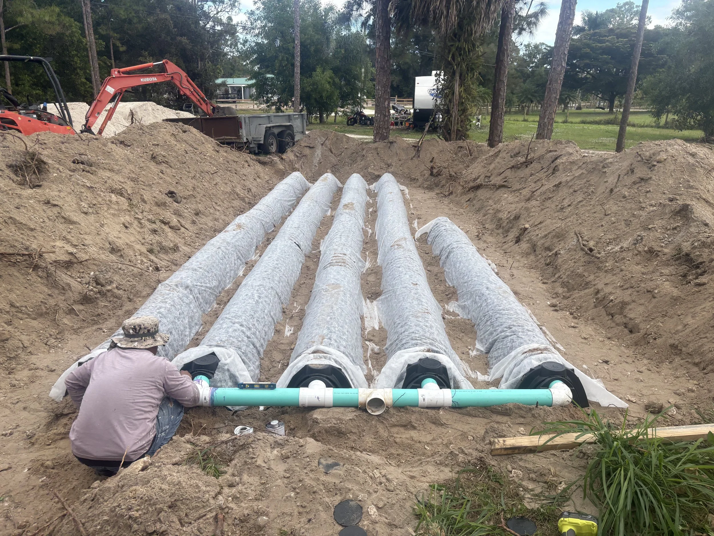 Workers installing large pipes underground using heavy equipment in a construction site surrounded by trees.