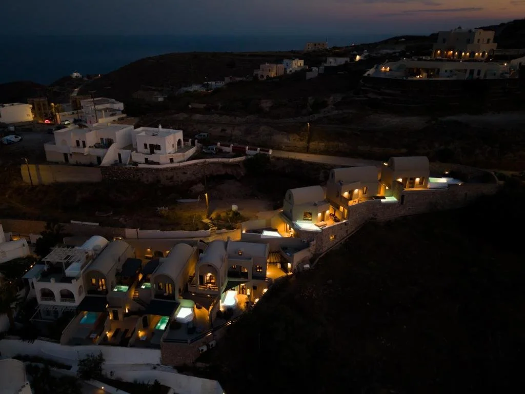 Nighttime view of white, cave-like houses with illuminated windows on a hillside near the ocean.