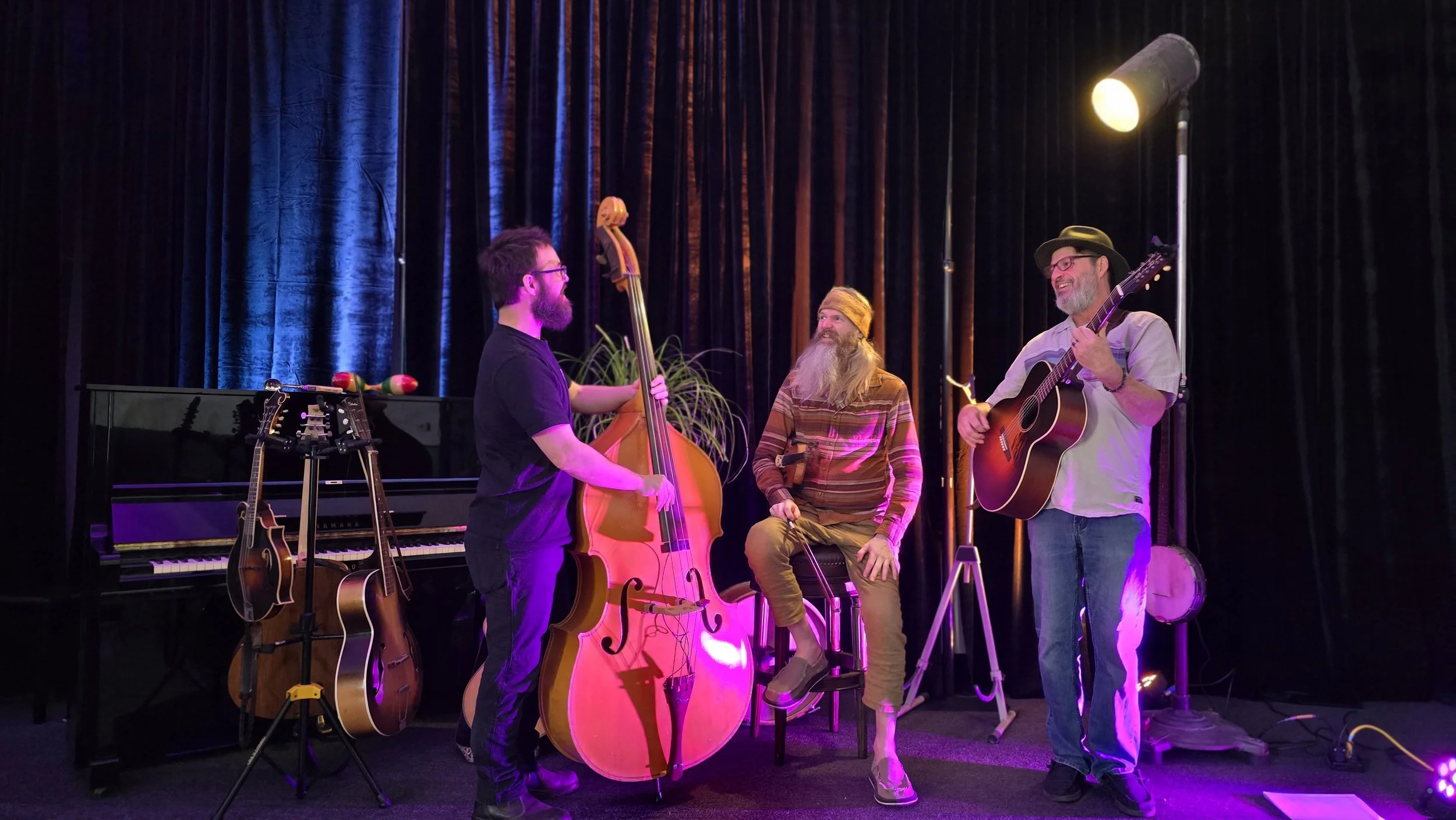Musicians performing live on stage; one plays upright bass, one plays guitar, one sits with percussion instruments, and one sings with a guitar; stage has beautiful black curtains, piano, and guitars.