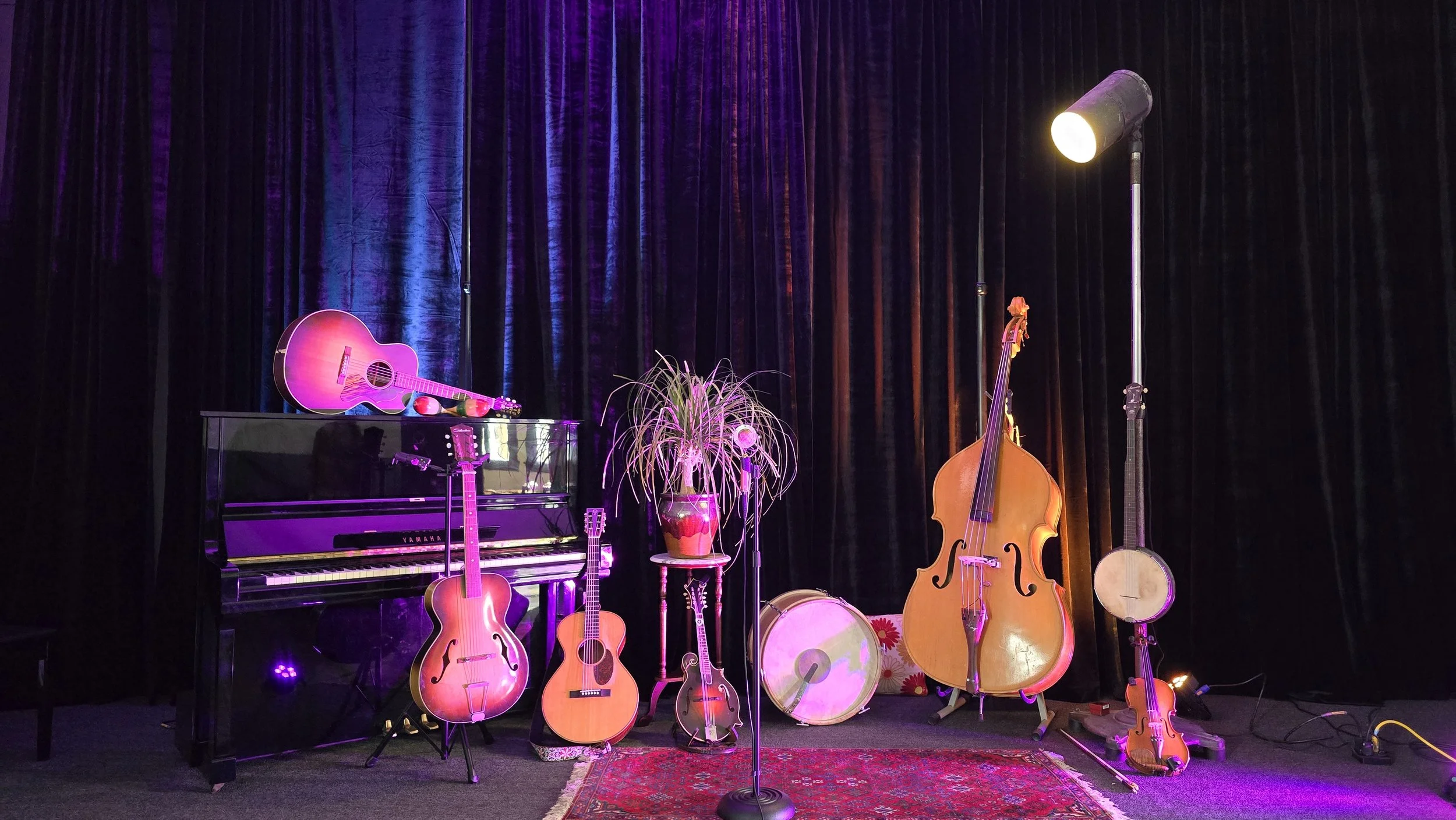 Stage with musical instruments including a pink guitar on a stand, a piano, an upright bass, several smaller guitars, a banjo, a tambourine, a floral-patterned drum, and a microphone, all set against dark purple velvet curtains, with stage lighting.