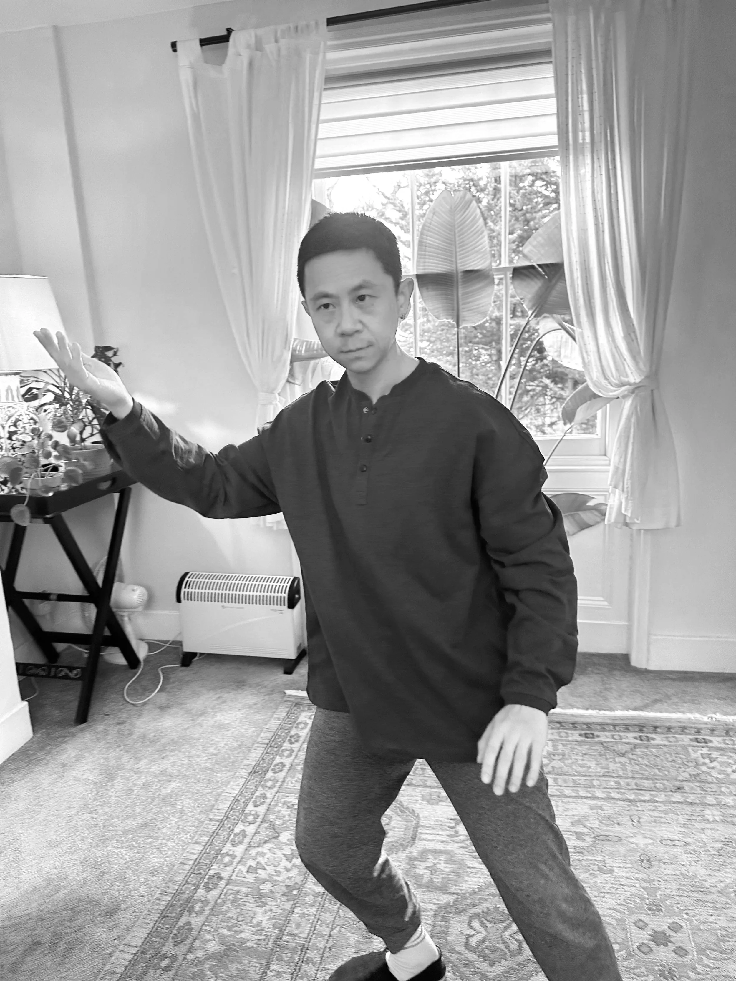 A man perform Tai Chi in a room with large windows, curtains, and plants, raising one arm. The room has a patterned rug and a heater on the floor.