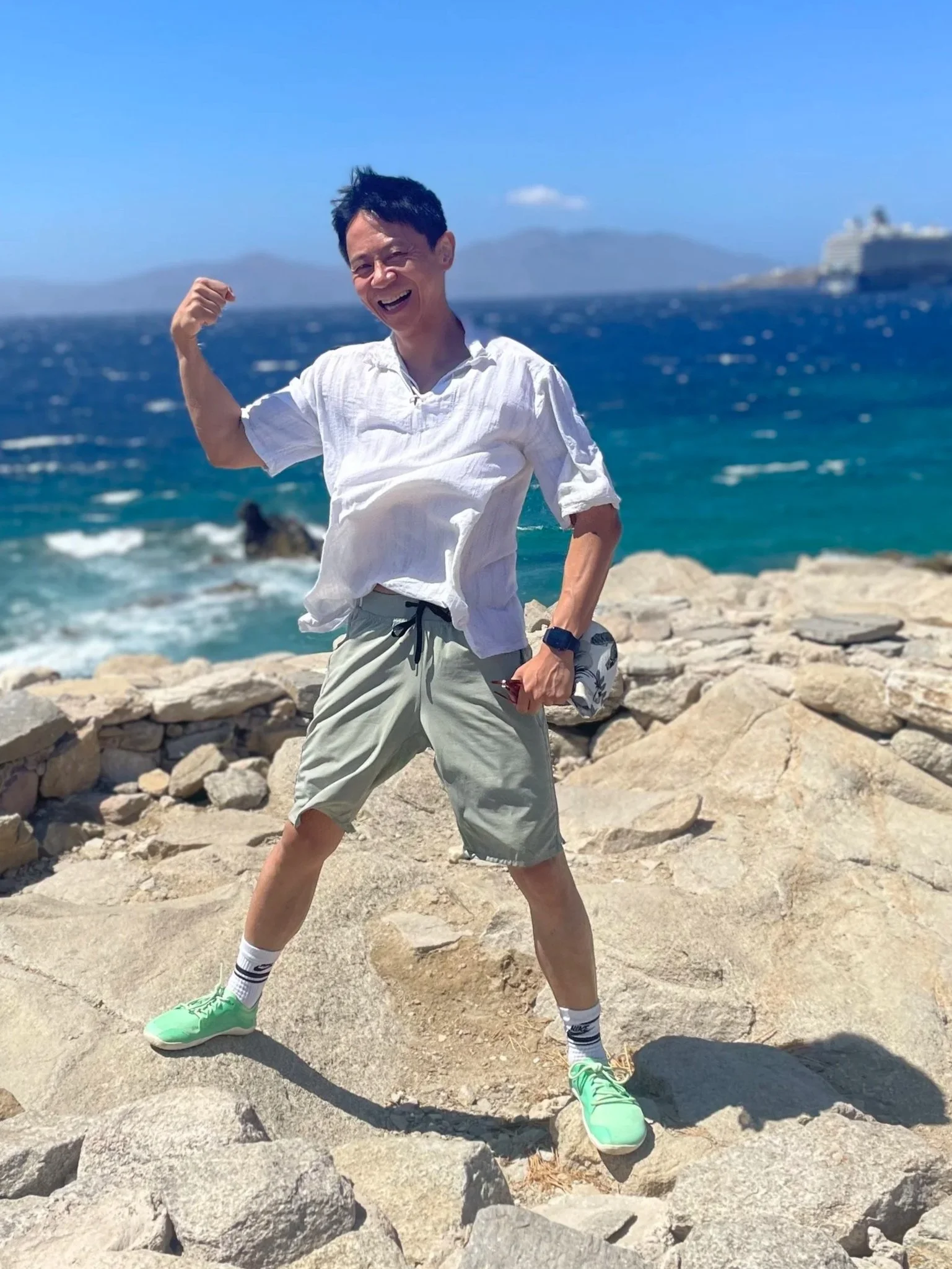 A man wearing a white shirt, shorts, and bright green sneakers pose with a flexed arm at a rocky beach with the ocean and a cruise ship in the background.