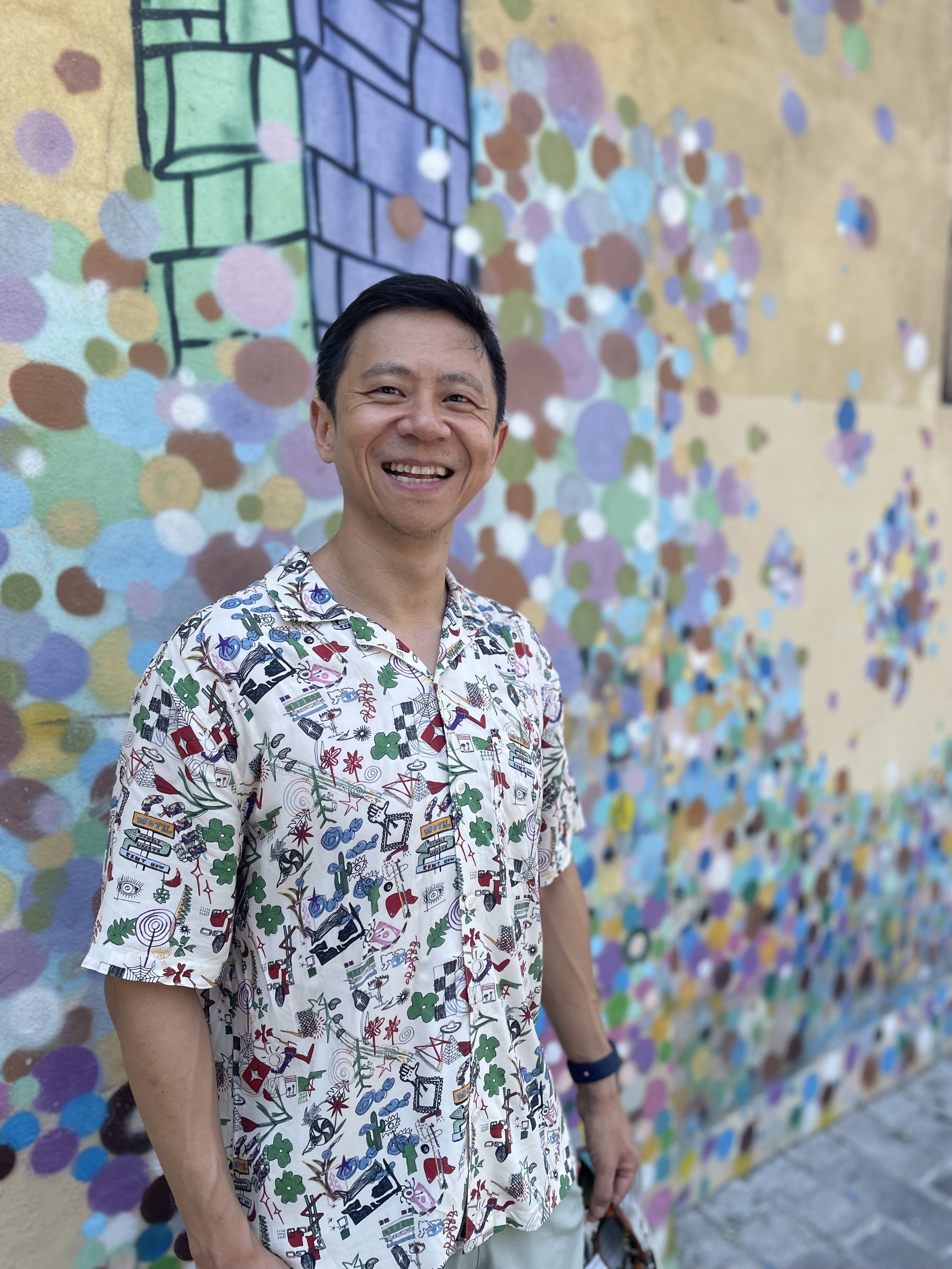 A smiling man standing in front of a colorful dot mural, wearing a white short-sleeve shirt with various colorful patterns.