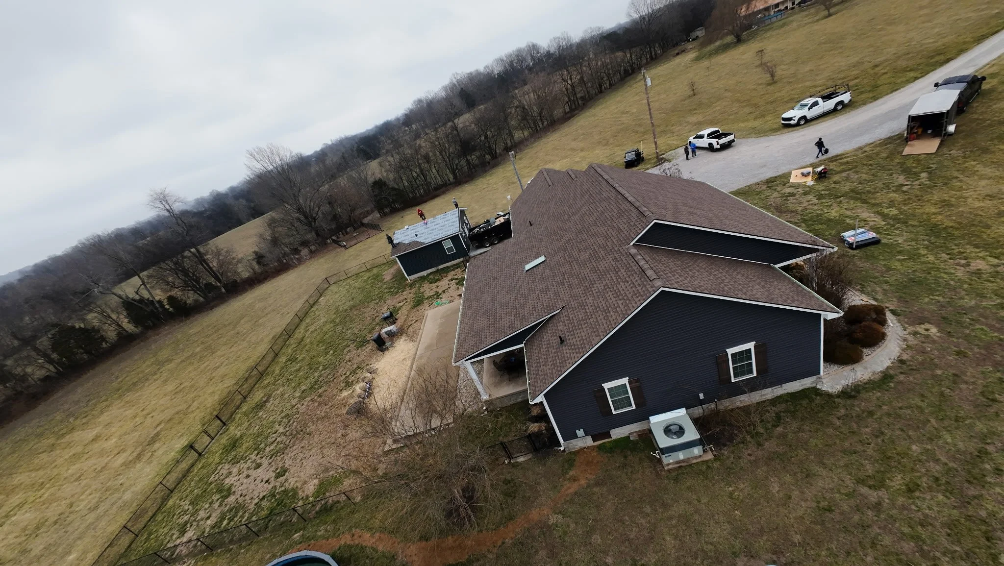 Aerial view of a black house with a brown roof in a rural setting, with a backyard enclosed by a fence, a patio, and a lawn. Several vehicles and equipment are parked nearby, and a few people are working or standing in the yard. The landscape include