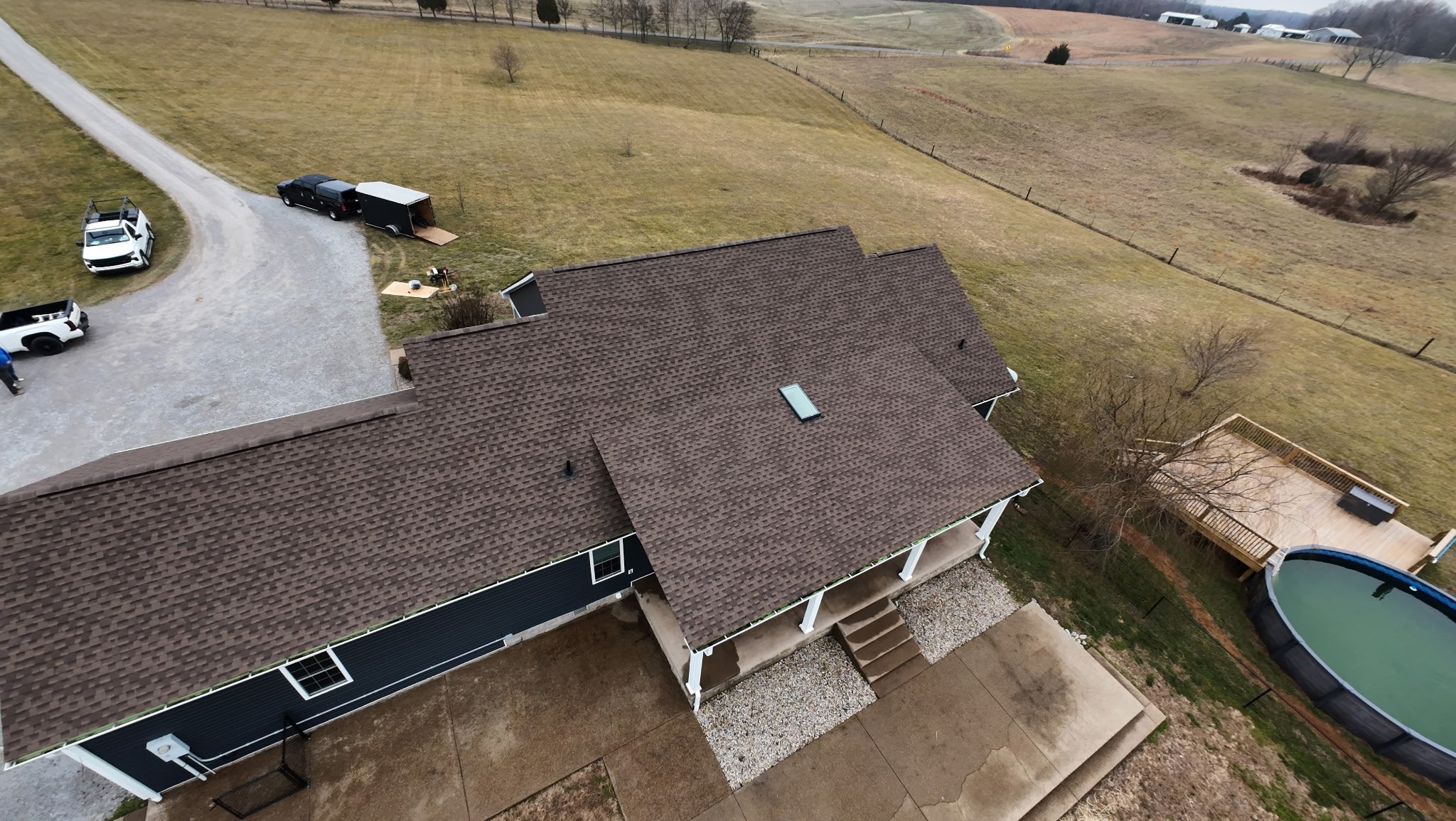 Aerial view of a backyard with a house, a small porch with steps, and a gravel patio. To the right, there is a deck with a tree and a fenced-in above-ground pool. In the background, open grass fields, a driveway with cars, and scattered trees are vis