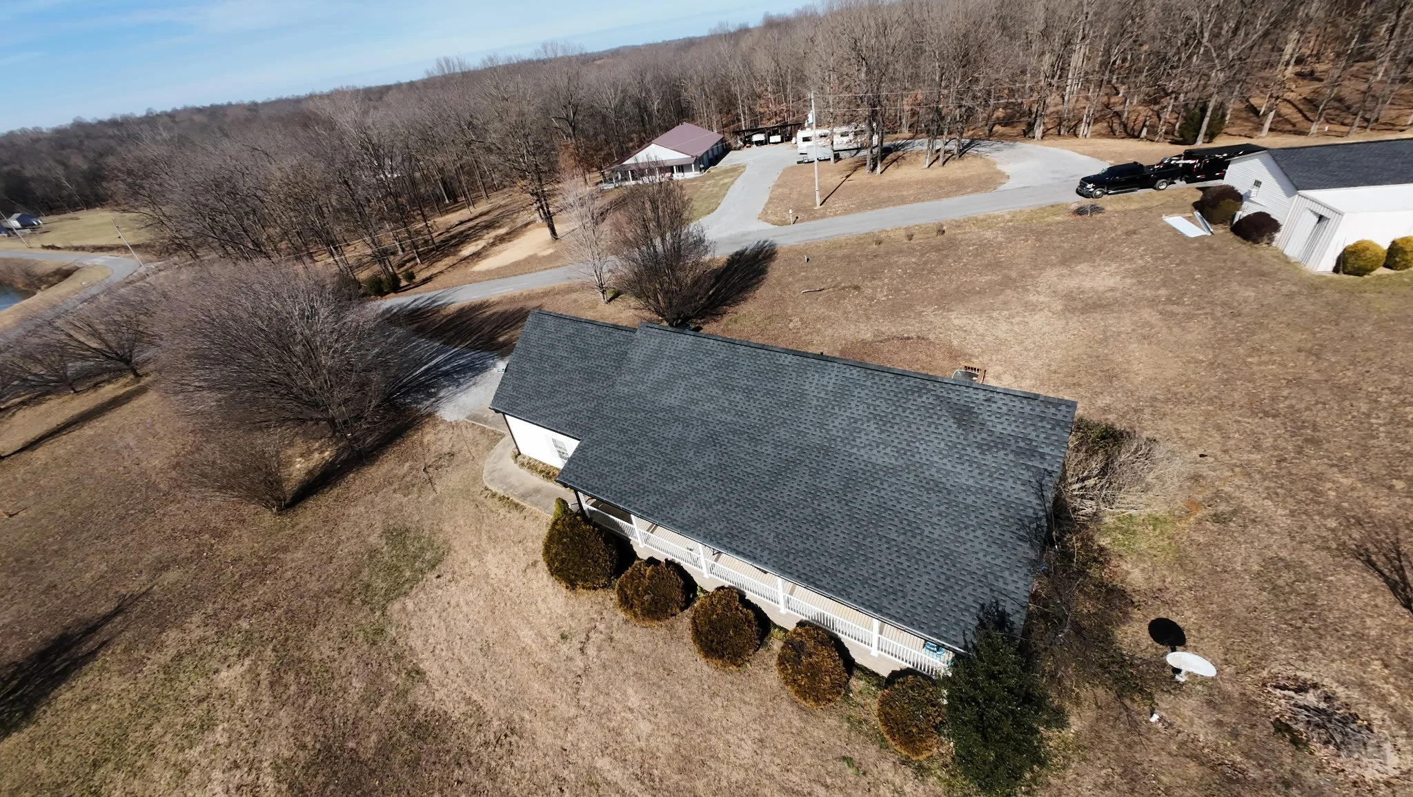 Aerial view of a residential yard with a house, leafless trees, a satellite dish, and nearby buildings, parking lot, and wooded area in the background.