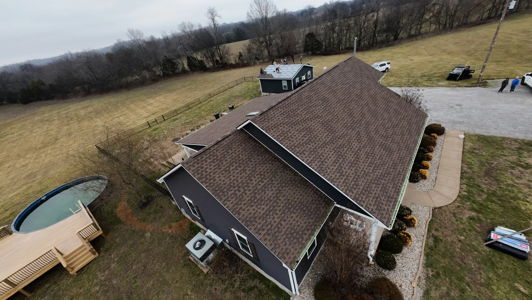 Aerial view of a house with a brown shingle roof, a concrete walkway, and a landscaped yard with bushes, a small pool, and a wooden deck with stairs. Several cars are parked on the driveway and grass field, with a group of people near the driveway. T
