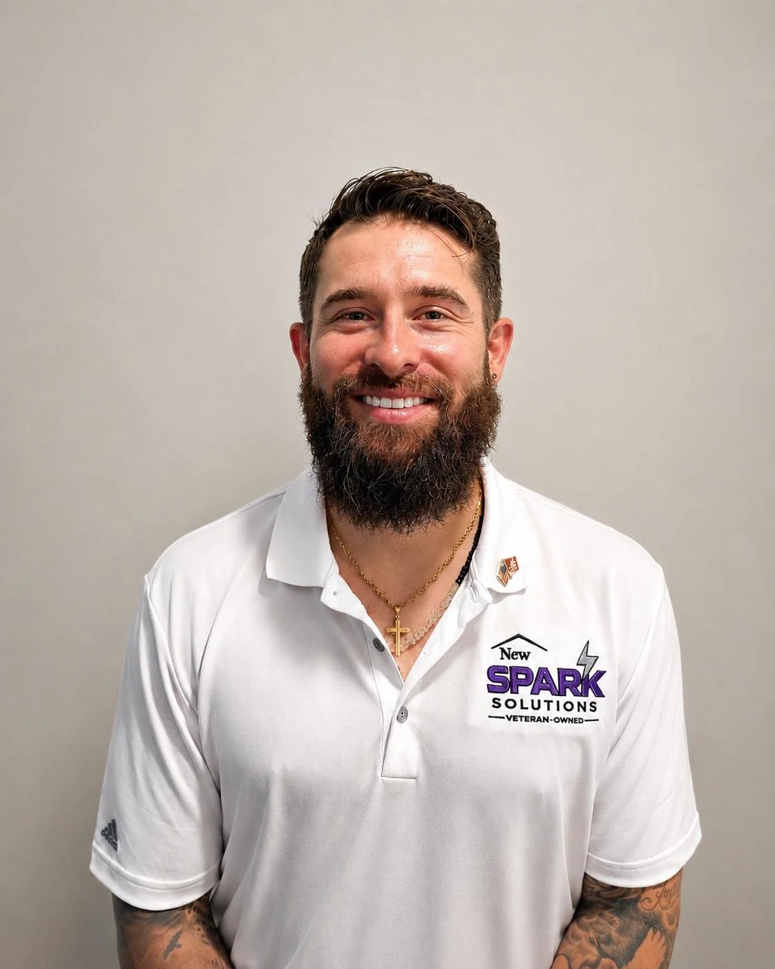 Man with a beard wearing a white polo shirt with the logo 'New Spark Solutions' and a pin, standing against a plain gray wall, smiling at the camera.