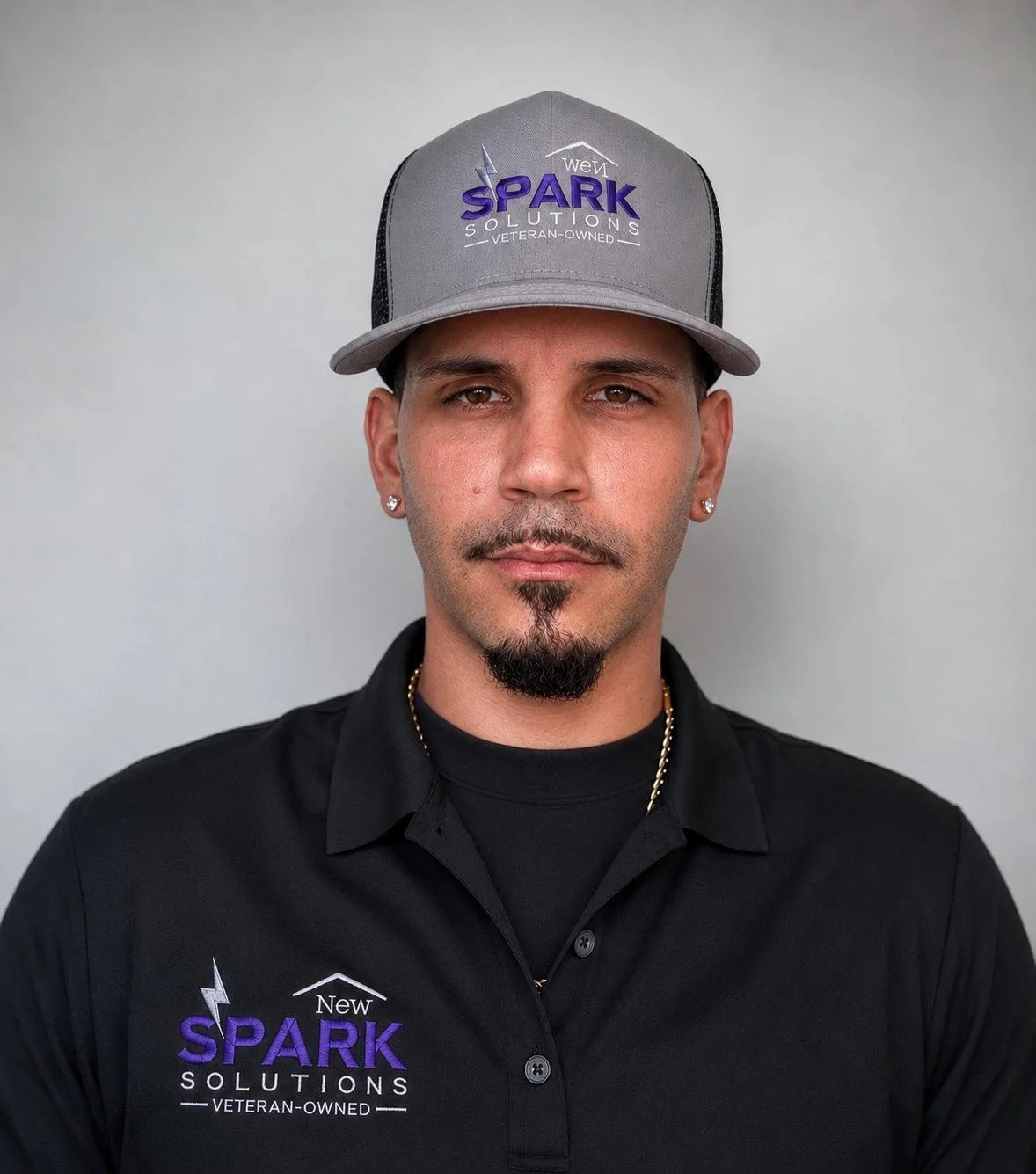 A man wearing a gray cap with the logo of Spark Solutions, a black shirt with the same logo, and earrings, standing against a plain gray background.