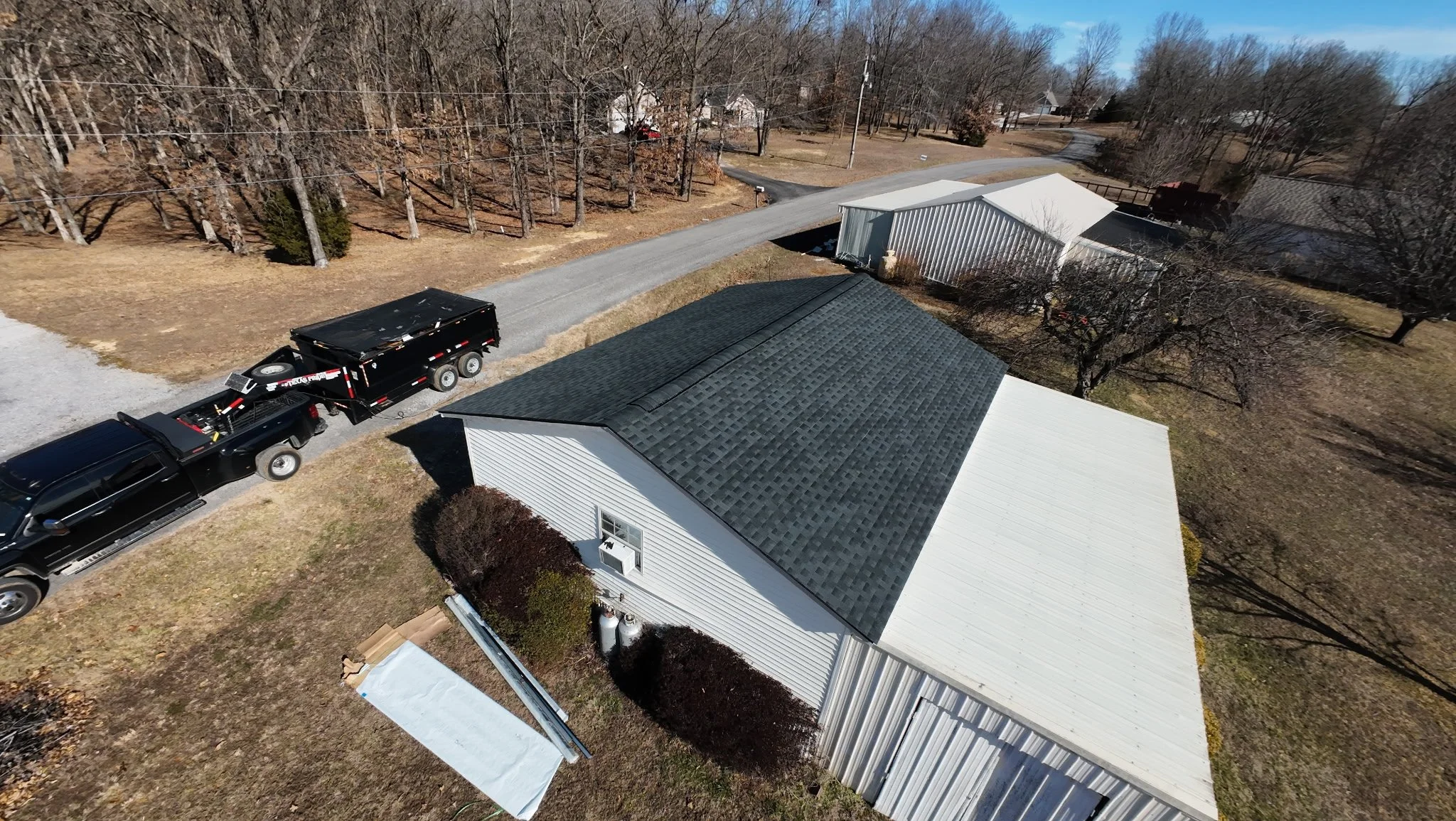 Aerial view of a house with a gray gabled roof, a white side wall, and small bushes at the front. A driveway runs alongside the house, with a black truck and a black trailer parked on it. Nearby, there is a large white metal building, leafless trees,