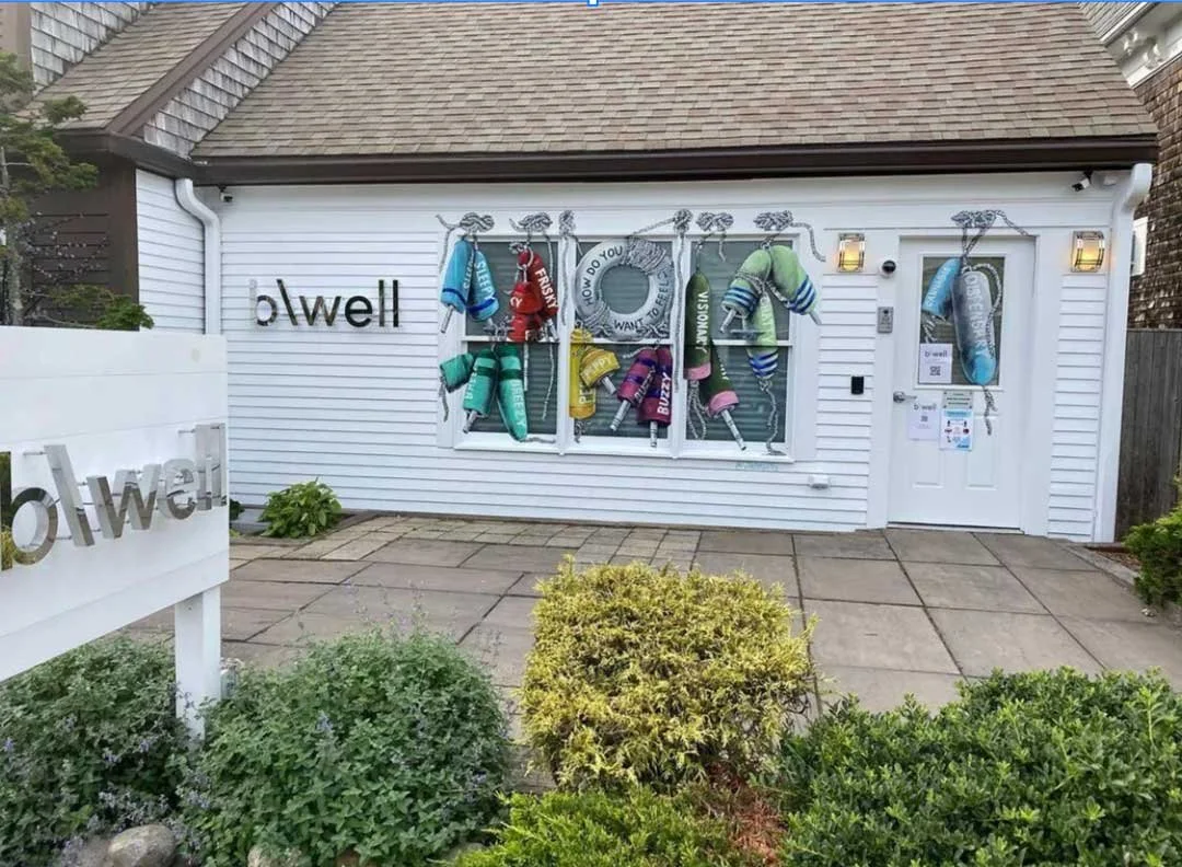The exterior of b\well cannabis dispensary in Provincetown, MA, featuring the b\well sign and a colorful mural of nautical buoys labeled with words like "Sleepy," "Frisky," "Happy," "Buzzy," and "Visionary"