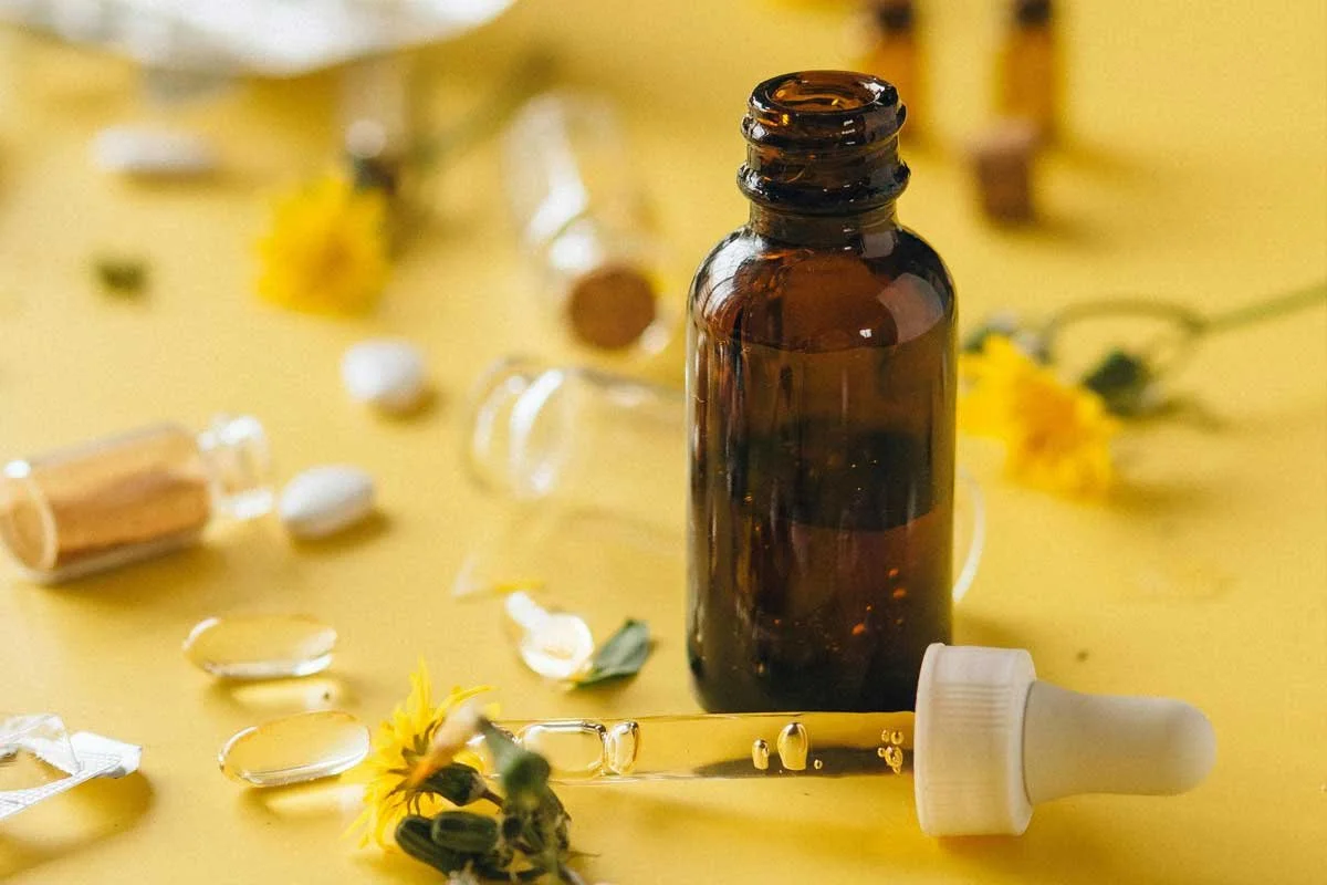 An amber dropper bottle of cannabis tincture with the dropper resting beside it, surrounded by yellow flowers, glass vials, and capsules on a bright yellow surface — available at b\well, Provincetown's queer owned cannabis dispensary.