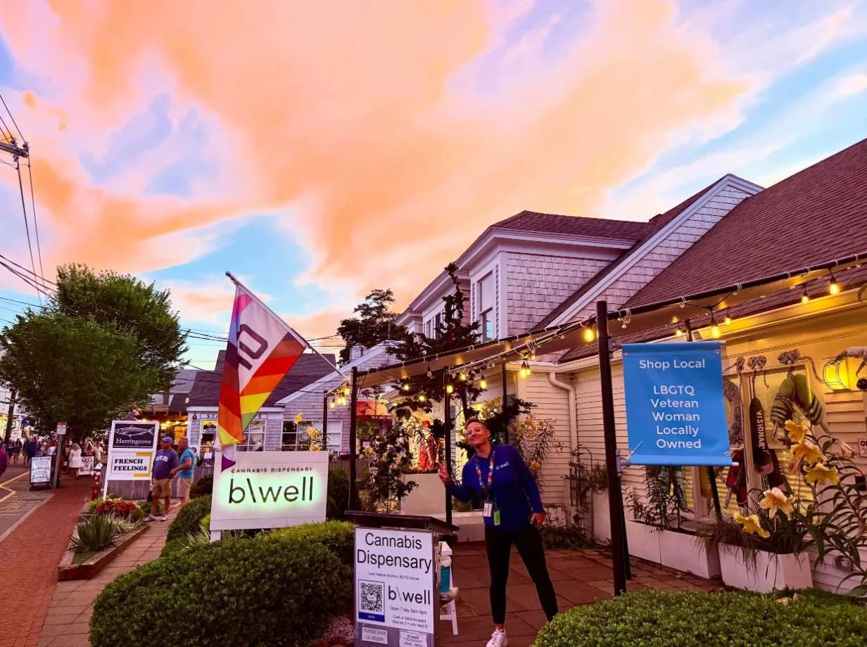 Woman standing in front of b\well dispensary storefront and sign at dusk, smiling at the camera