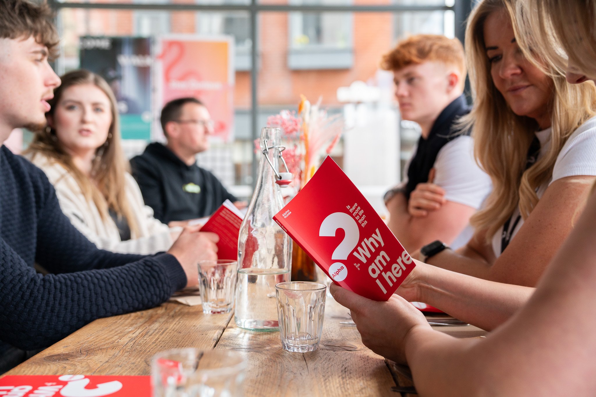A group of young adults sitting at a wooden table during a discussion or meeting, with a person holding a red pamphlet titled "Who am I here" and an alphabet logo on it. The setting appears to be a casual indoor space with large windows.