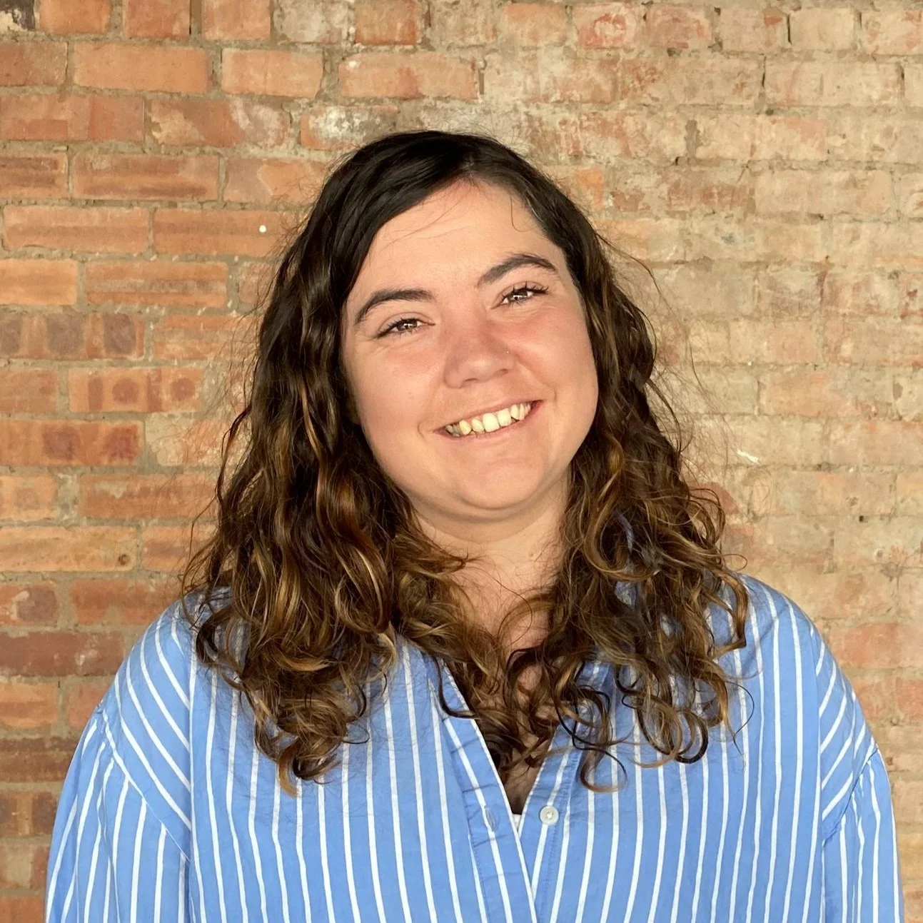 A woman with curly brown hair smiling in front of a brick wall, wearing a blue and white striped shirt.