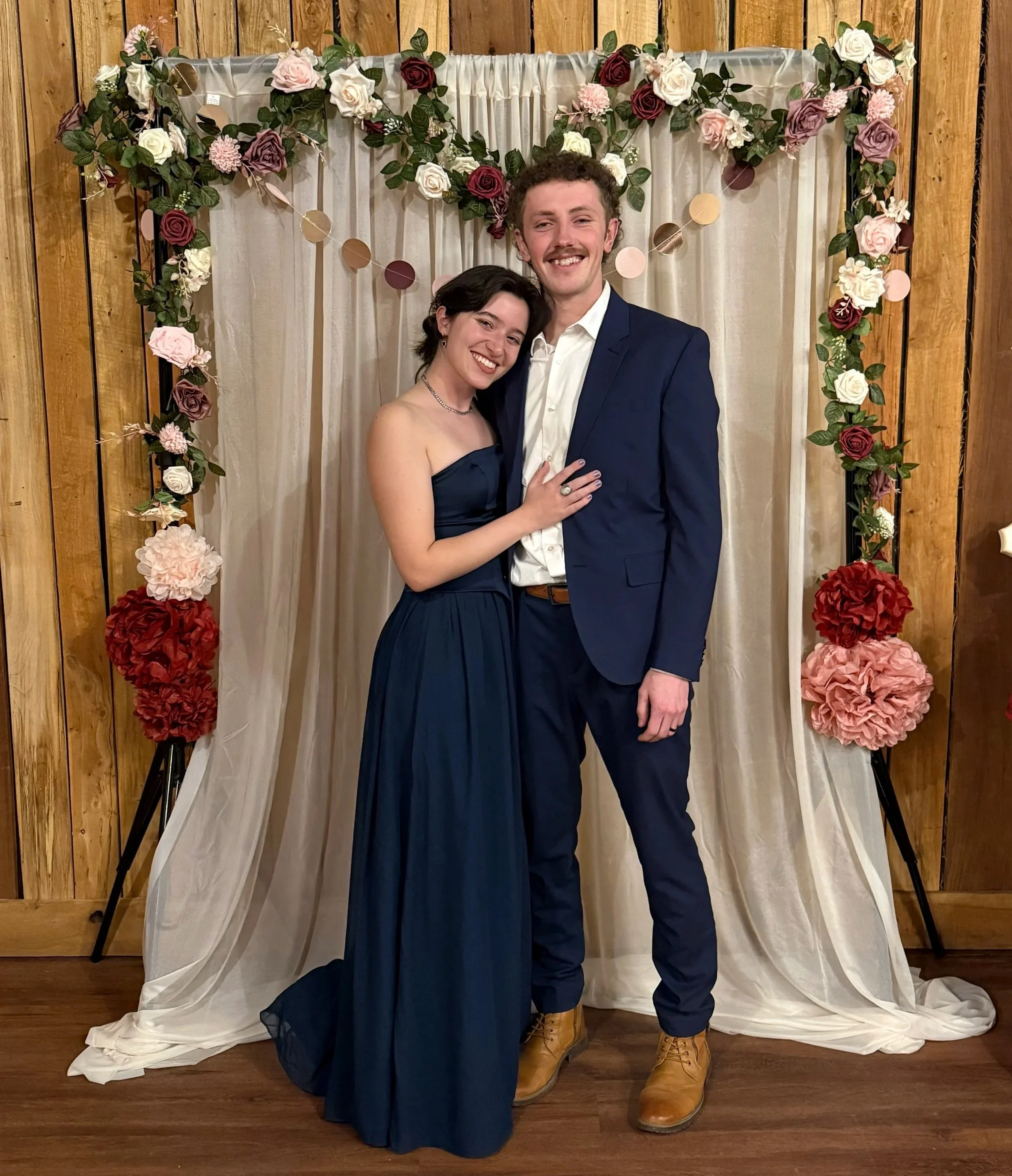 A young woman and a young man stand close together, smiling, in front of a floral backdrop with pink, red, and white flowers and cream-colored drapes. The woman is wearing a strapless navy blue gown, and the man is dressed in a navy blue suit with a white shirt and tan boots. They appear to be at a formal event or celebration.