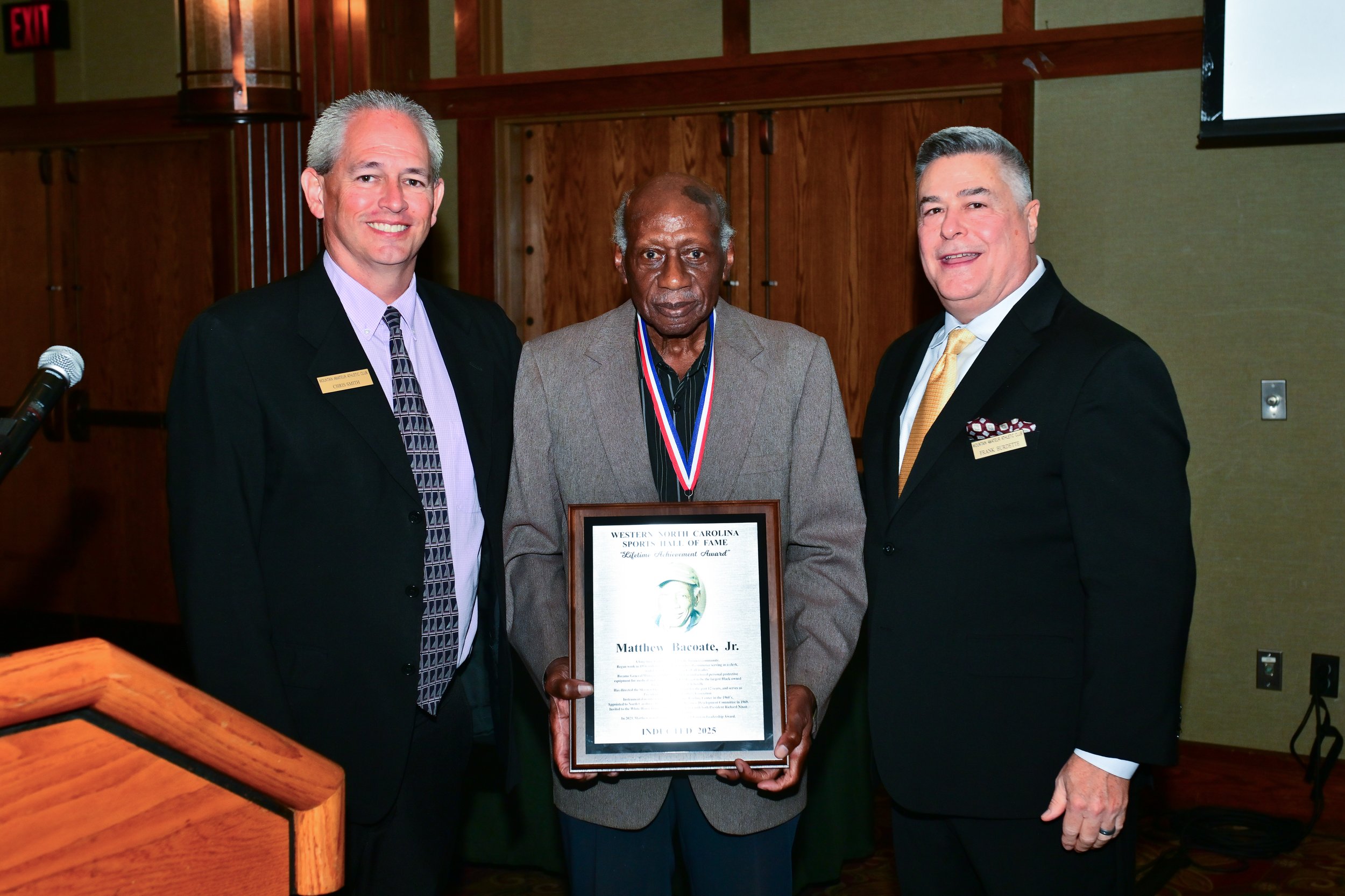 Three men standing together at an award ceremony. The man in the middle holds a framed certificate and wears a medal around his neck. The men on the sides are dressed in suits, and the background includes a wooden wall and a screen.