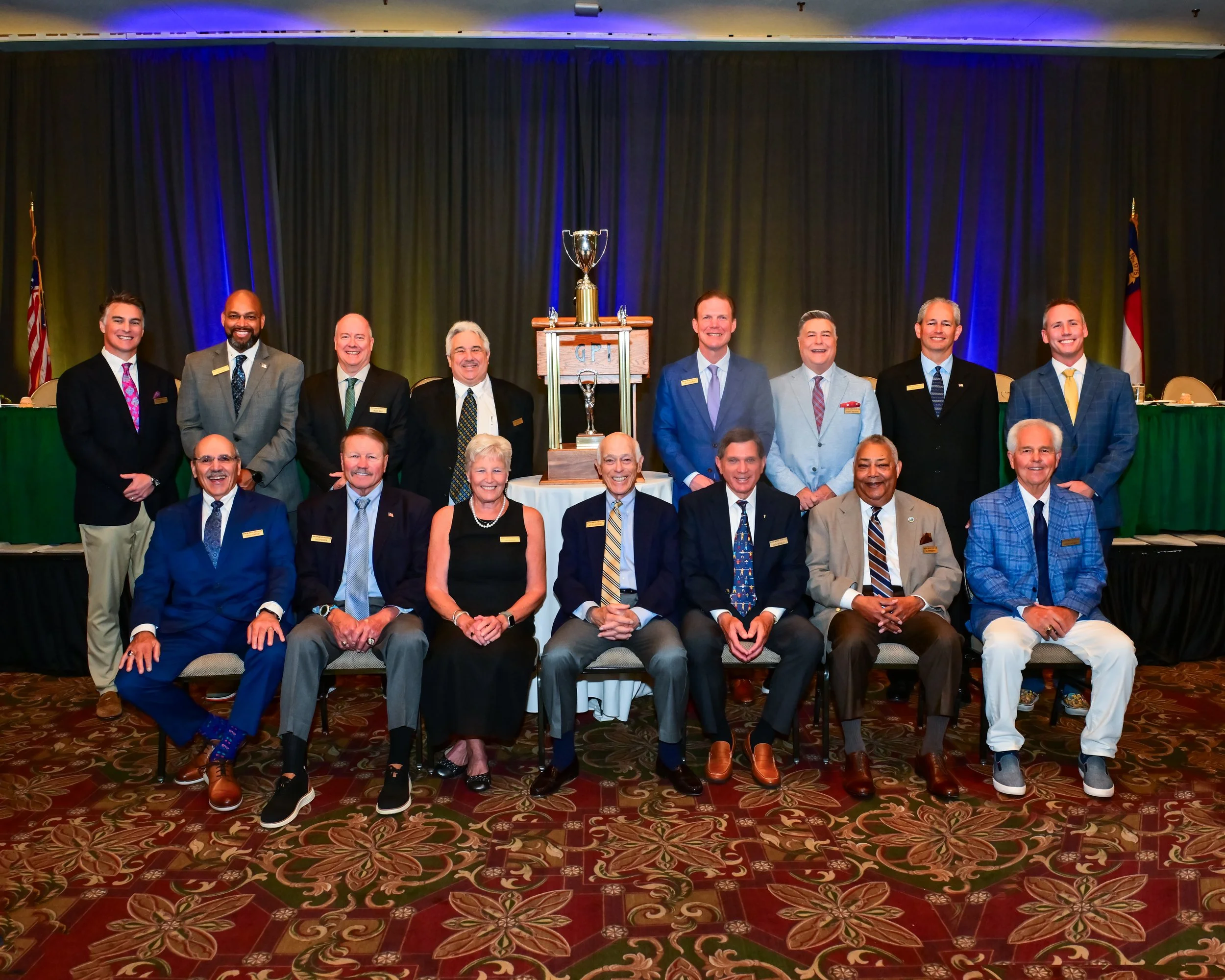 Group of people in formal attire standing and sitting in front of a black curtain, with a trophy on a stand in the center of the group, at an indoor event.